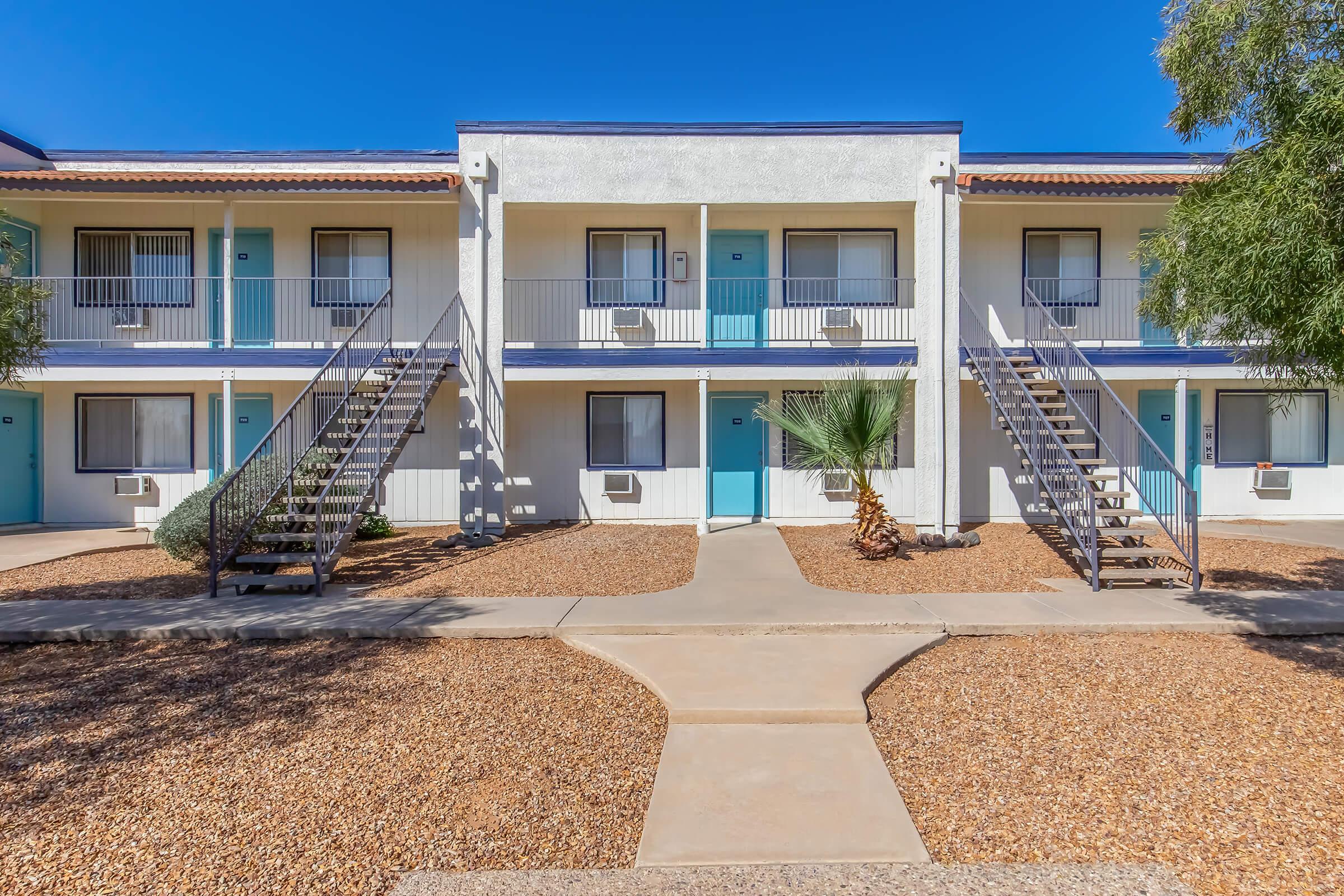 Exterior view of a two-story apartment building with a light-colored facade. It features staircases on both sides leading to individual units, each with turquoise doors and small air conditioning units. The surrounding area has landscaped gravel with a few plants, under a clear blue sky.