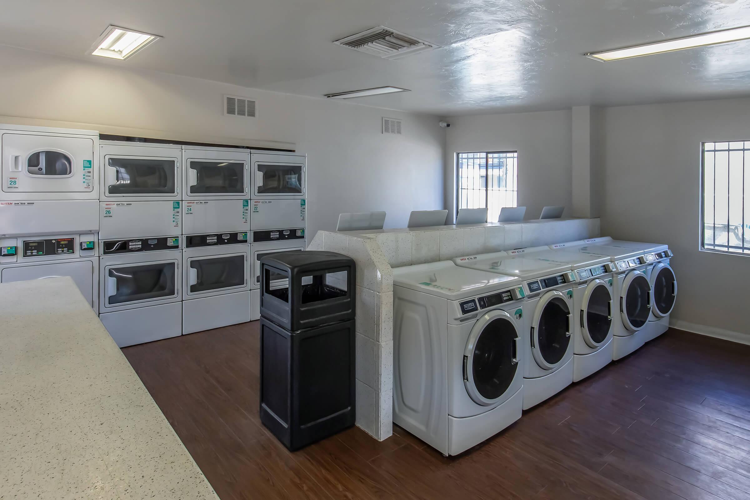 A clean and well-lit laundry room featuring multiple washers and dryers lined up along the wall, a counter area, and a black trash bin. There are windows allowing natural light, and folding tables can be seen in the background, creating a functional space for laundry tasks.