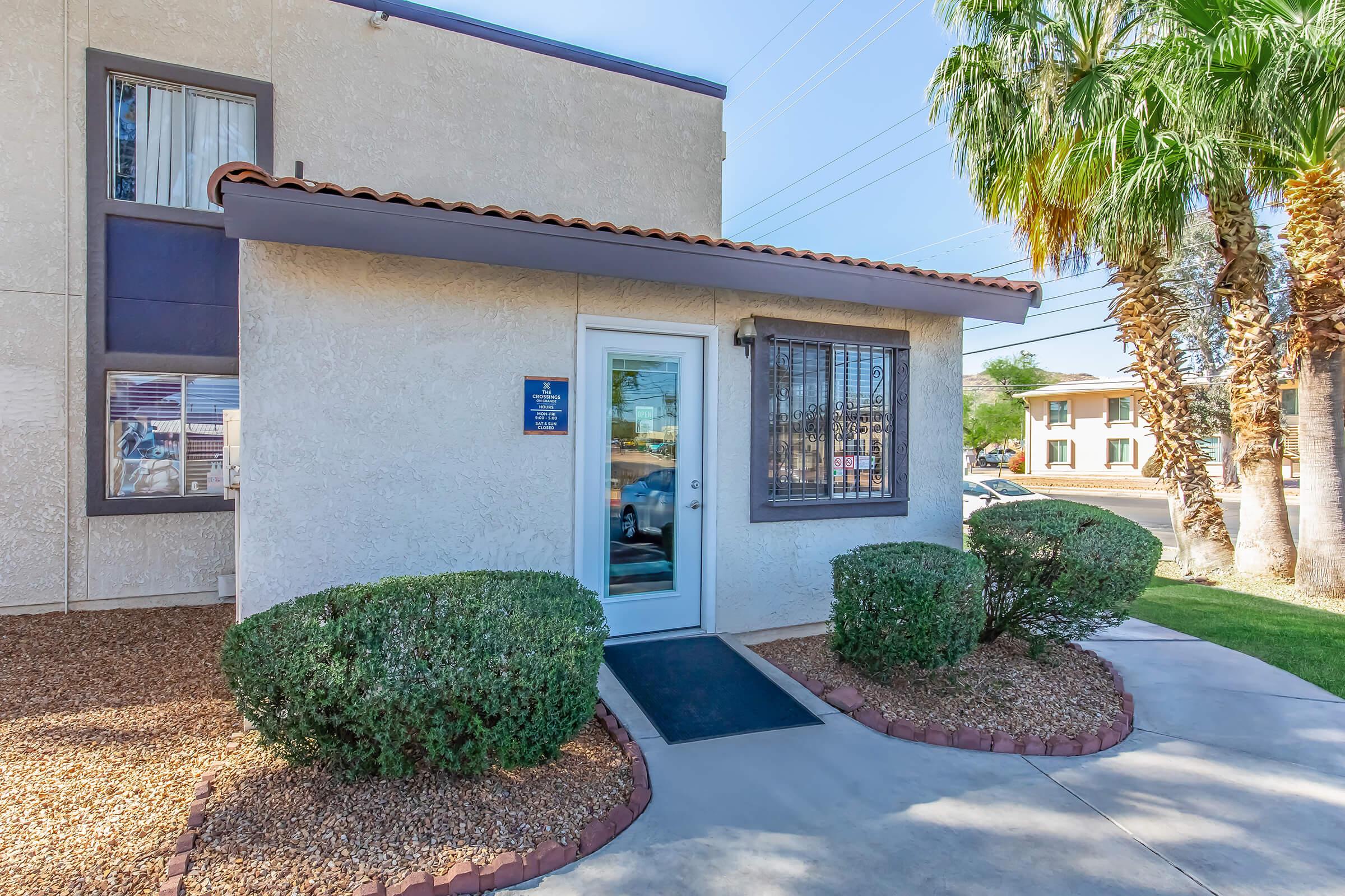 Exterior view of a building entrance featuring a white door and a window with decorative bars. The surrounding area has well-maintained shrubs and palm trees, and a paved pathway leads to the door. Clear blue skies are visible in the background.
