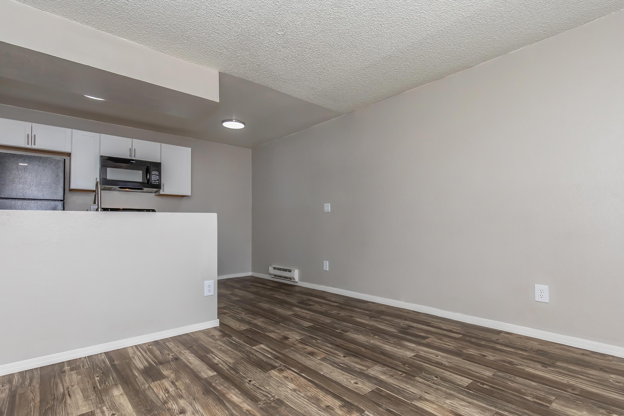A modern, spacious living area featuring light-colored walls and hardwood-style flooring. An open kitchen area is visible, with white cabinetry and appliances. Soft natural light fills the room, and there's an air conditioning unit on the wall, contributing to a clean and inviting atmosphere.