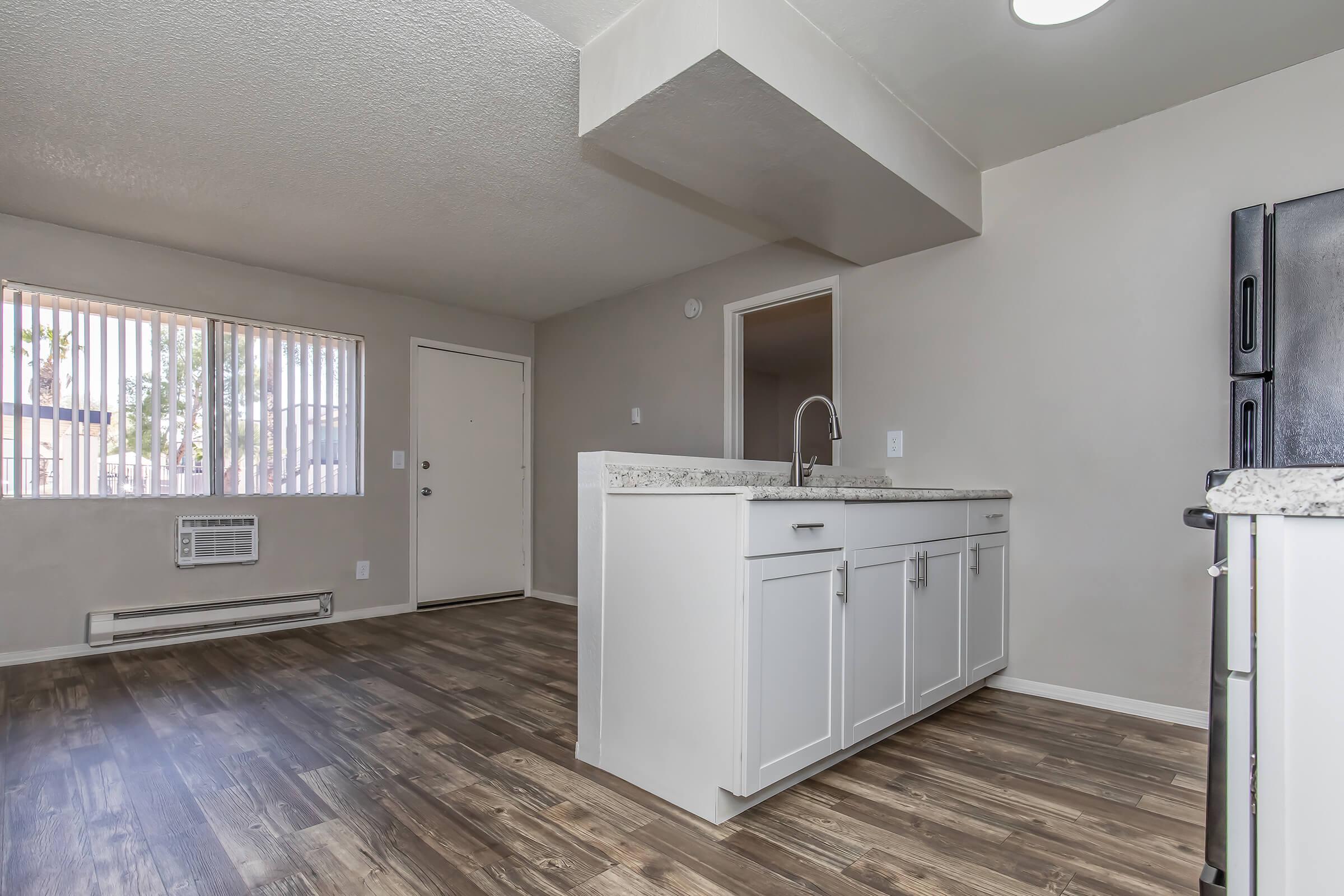 Interior view of a modern kitchen and living area. The kitchen features a granite countertop with a sink, white cabinetry, and a black refrigerator. The room has wood-like flooring, a wall-mounted air conditioner, and a door leading outside. Large windows provide natural light.
