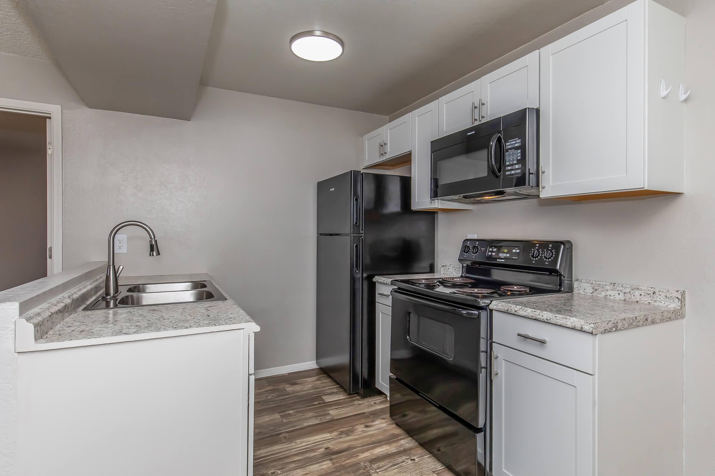 Modern kitchen featuring white cabinetry, black appliances including a refrigerator, microwave, and stove. The countertops are speckled granite, and there is a double sink with a high-arc faucet. Natural light is provided by a ceiling fixture, and the flooring is wood-like laminate.