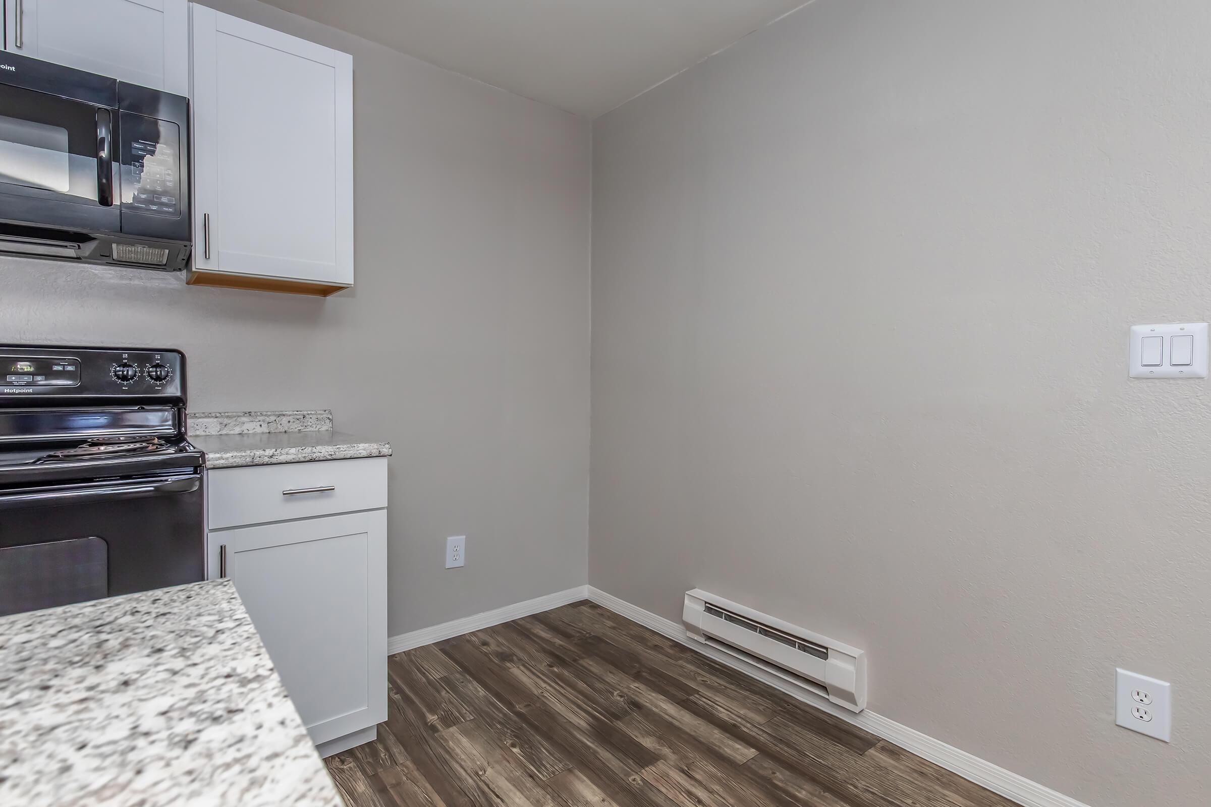 A modern kitchen featuring a black stove and microwave, white cabinets, and granite countertops. The flooring is wood-like, and the walls are painted a light gray. There is a heater unit on the wall, and the space is well-lit and uncluttered.
