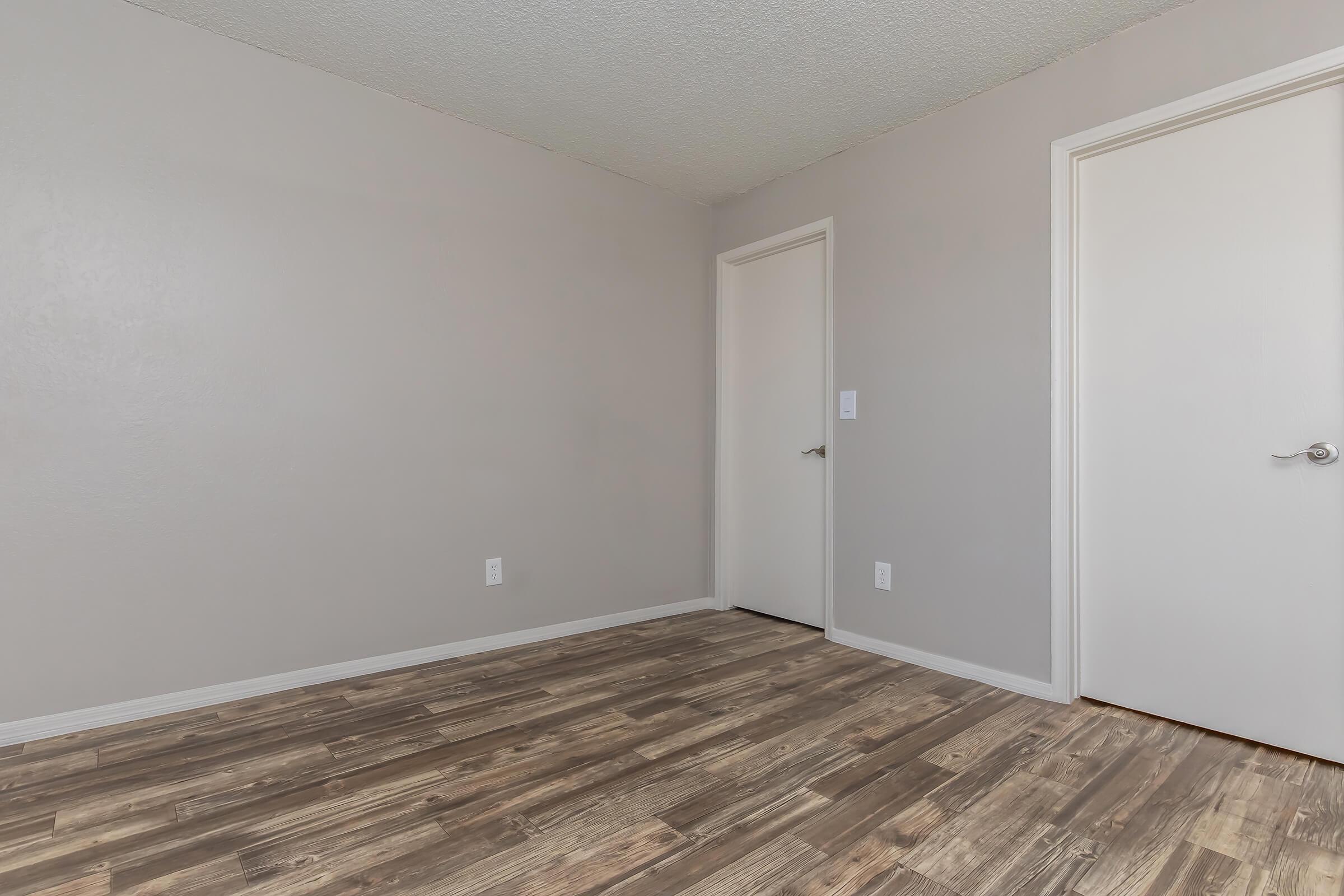 A simple, empty room with light gray walls and wooden flooring. There are two closed white doors on opposite walls, and the overall space appears neat and unadorned, suitable for various purposes such as a bedroom or office.