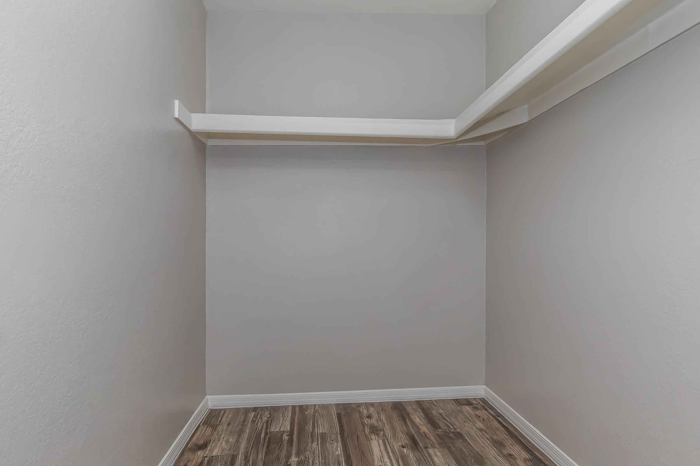 A vacant closet with light gray walls and a wooden floor. Two white shelving units are mounted on the walls, providing storage space. The overall ambiance is clean and minimalistic, with no items present in the closet, giving a spacious appearance.