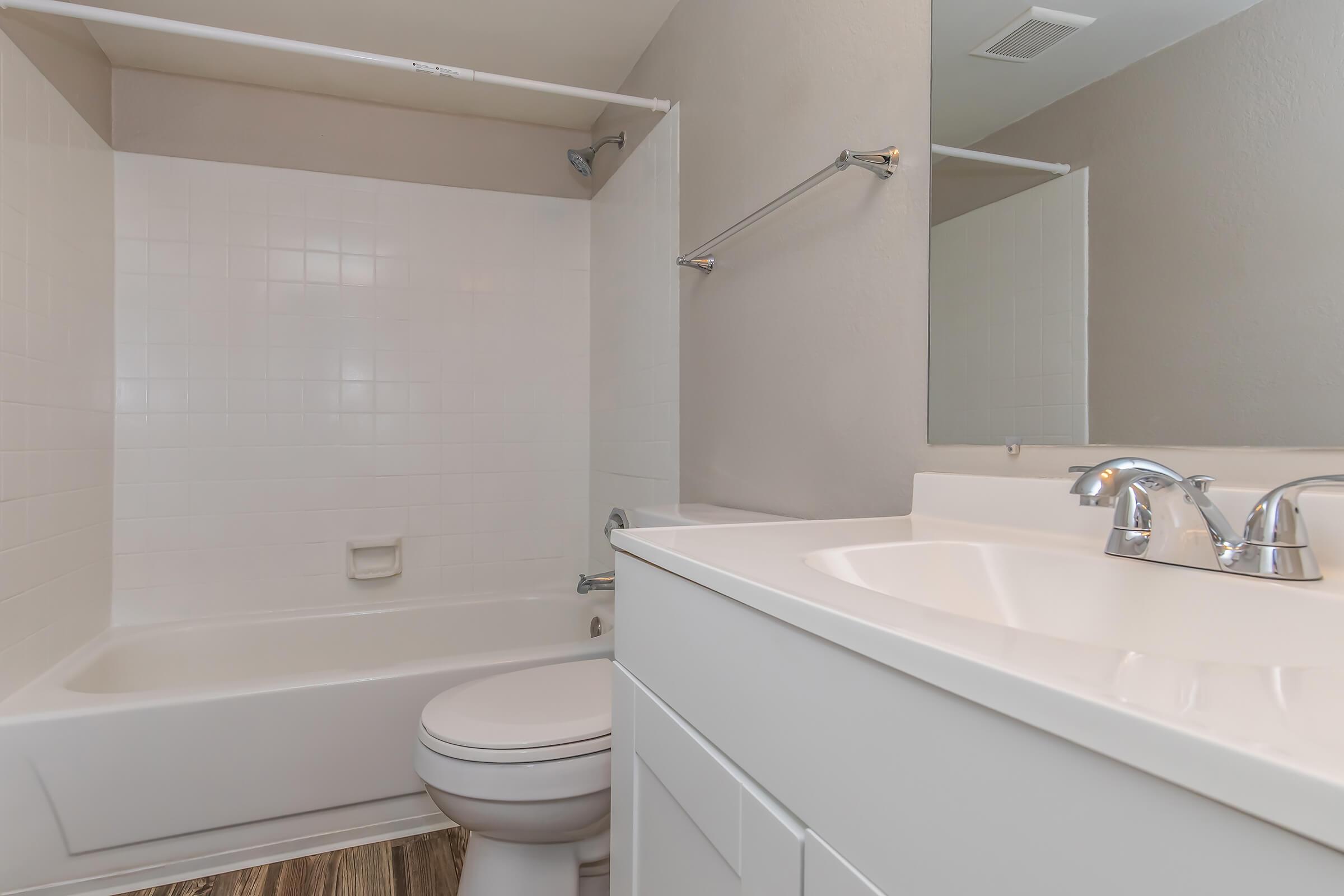 Bright and clean bathroom featuring a white bathtub and shower combination, a modern vanity with dual faucets, and a large mirror. The walls are painted in a soft neutral tone, and the flooring appears to be a wood-like material, giving the space a warm and inviting feel.