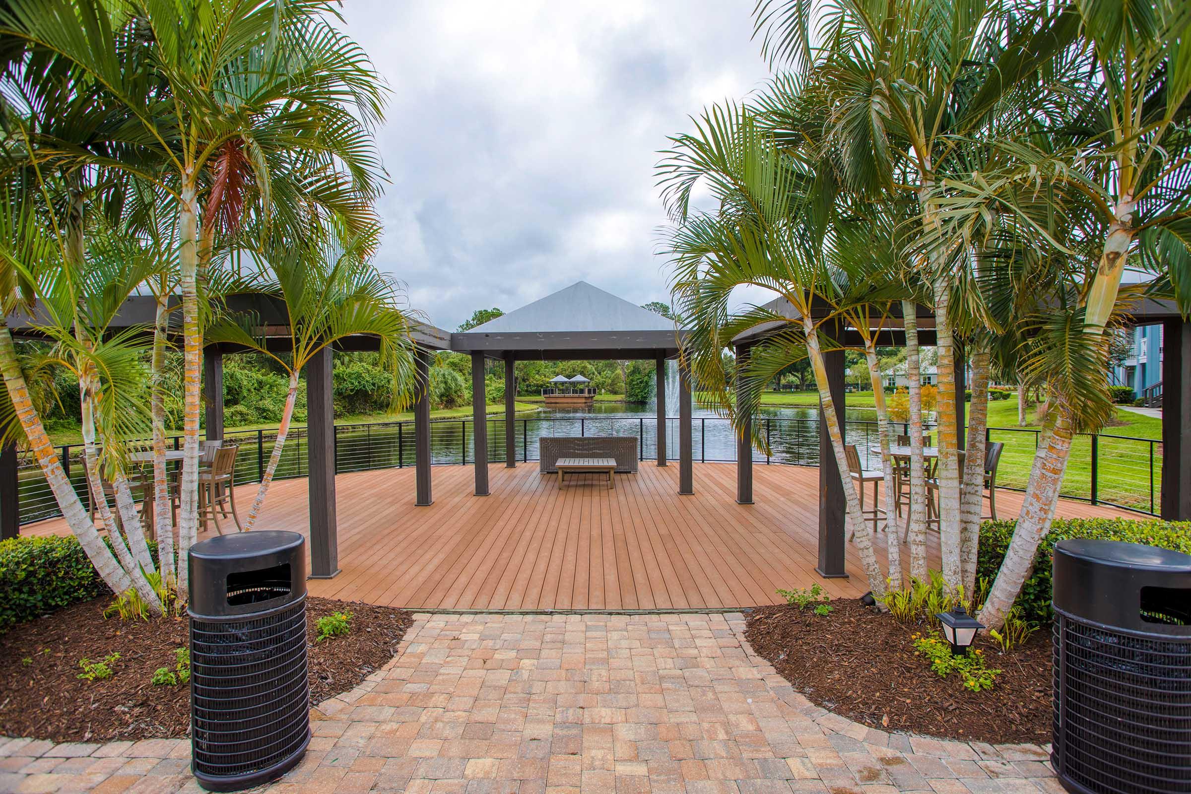 A scenic outdoor area featuring a wooden deck surrounded by palm trees, two pavilions with seating, and a view of a serene lake. The sky is partly cloudy, and landscaped areas with mulch and potted plants enhance the inviting atmosphere.