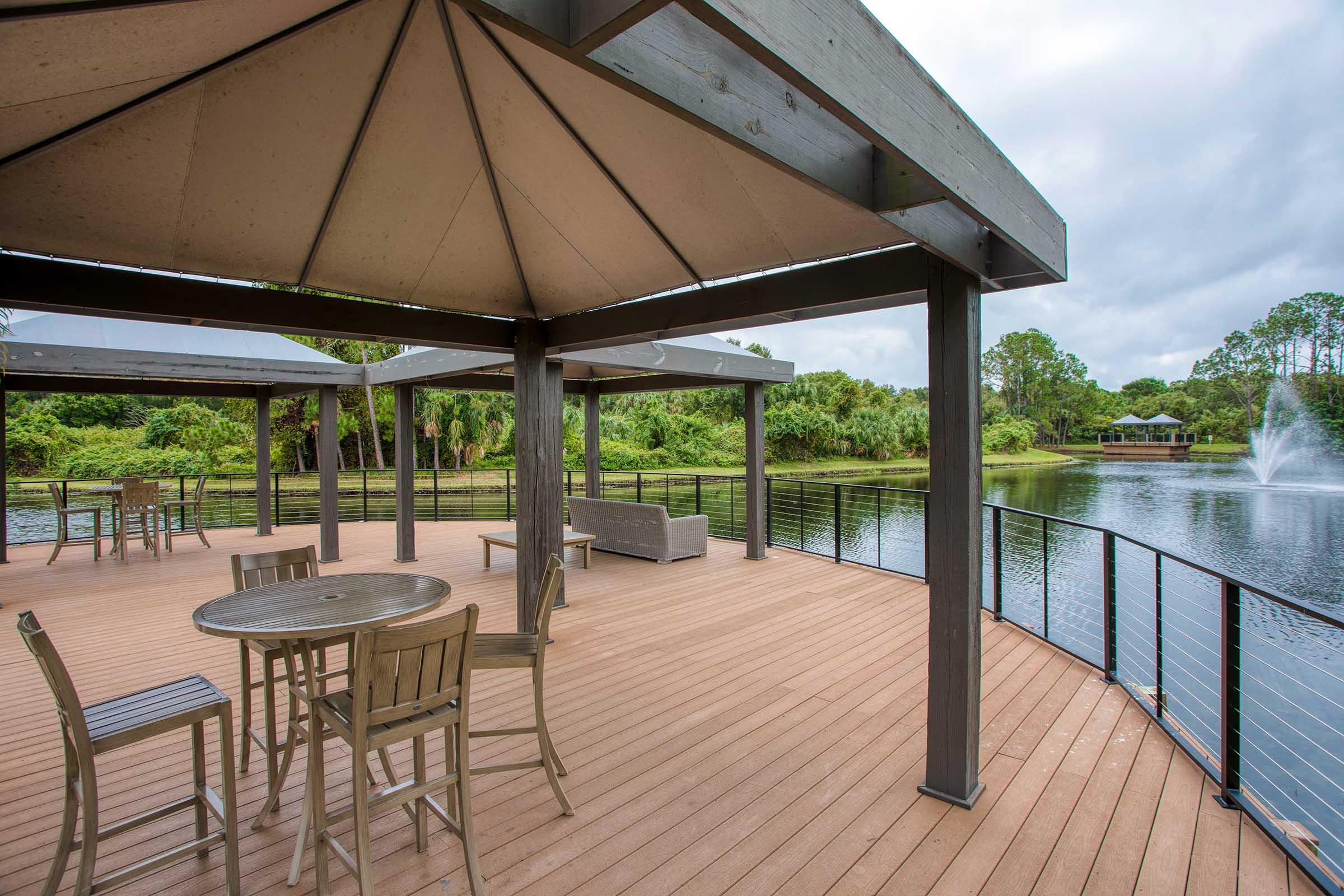 A wooden deck overlooking a serene lake, featuring several shaded seating areas with tables and chairs. Lush greenery surrounds the water, and a fountain can be seen in the distance. The sky is partly cloudy, creating a calm and inviting atmosphere.