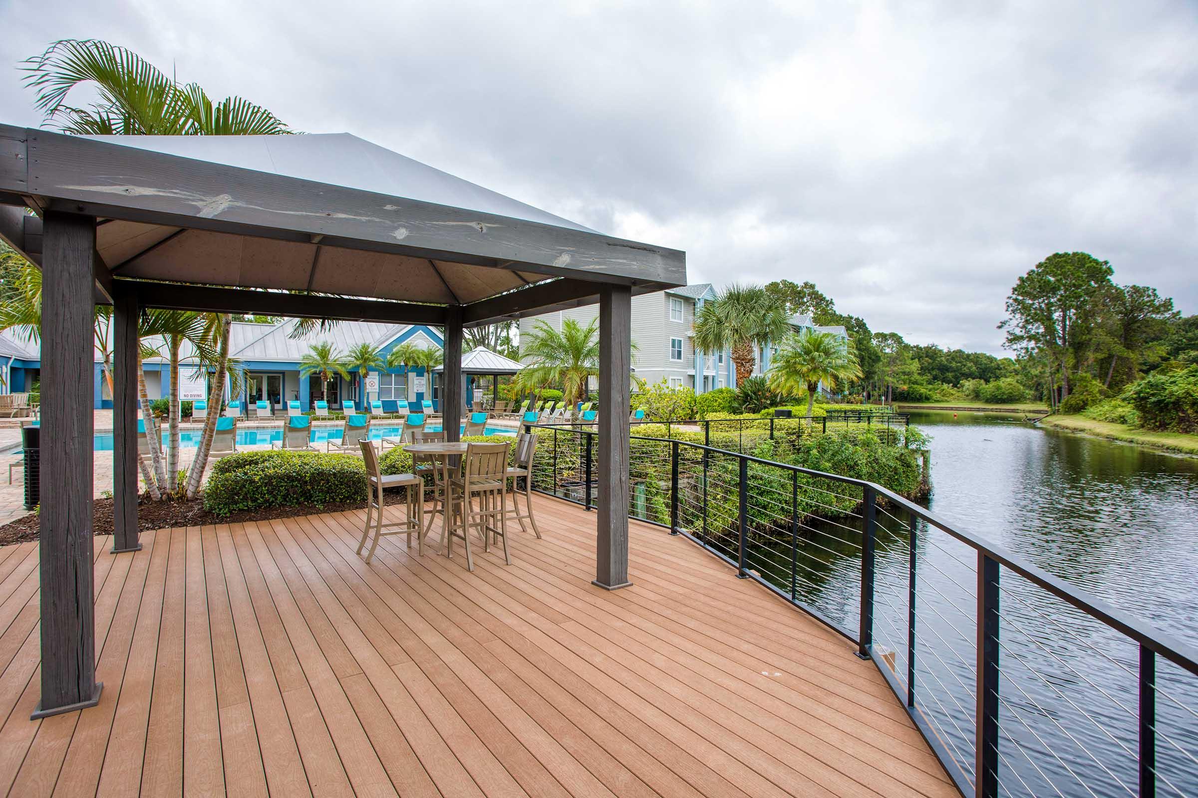 A wooden deck with a gazebo overlooking a tranquil water body. In the background, a pool and palm trees are visible, along with buildings. The sky is overcast, creating a calm atmosphere.