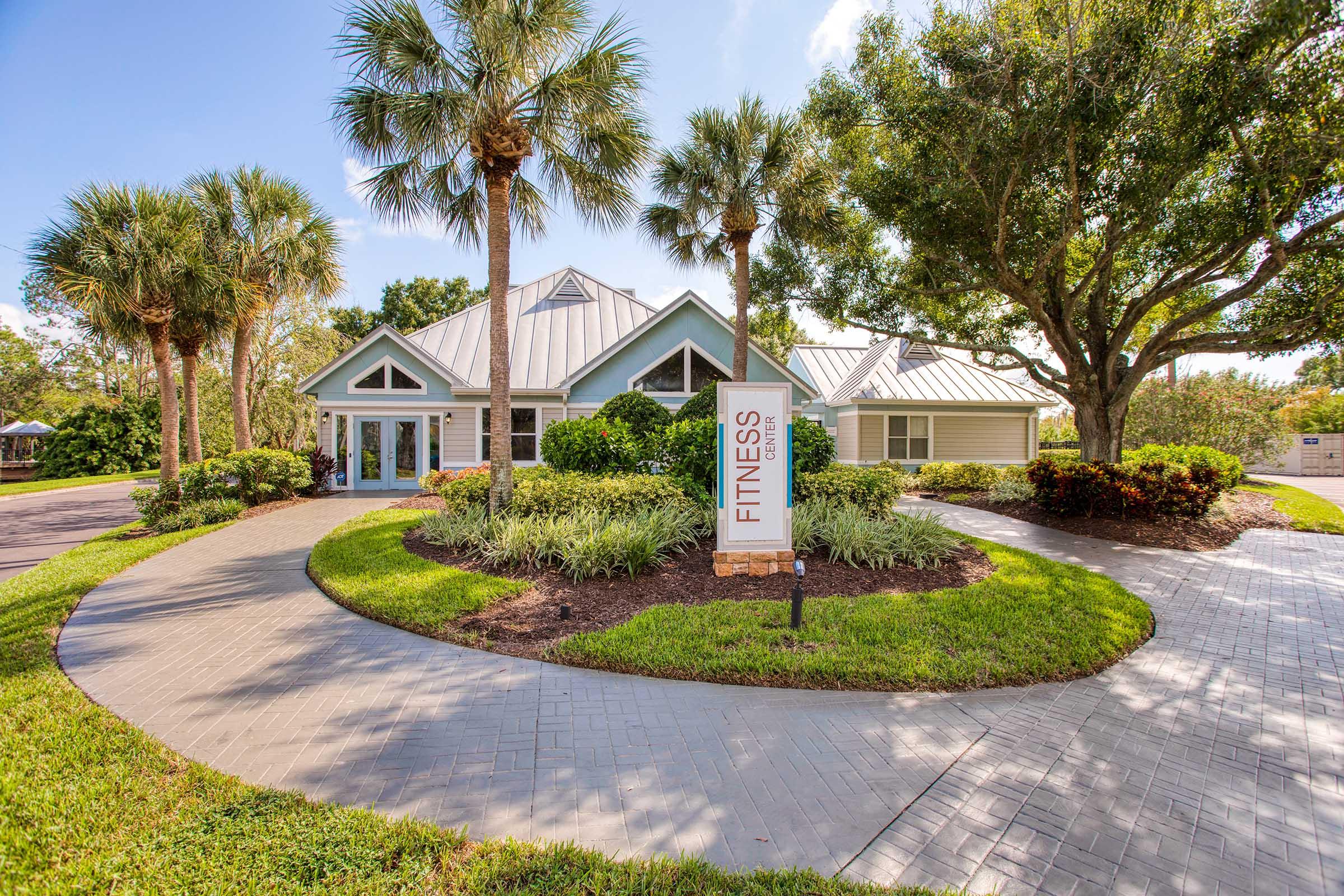 A modern fitness center building surrounded by manicured landscaping, palm trees, and green shrubs. A pathway leads to the entrance, and a sign indicates the facility's purpose. The setting is bright and inviting, showcasing a well-maintained exterior in a residential area.