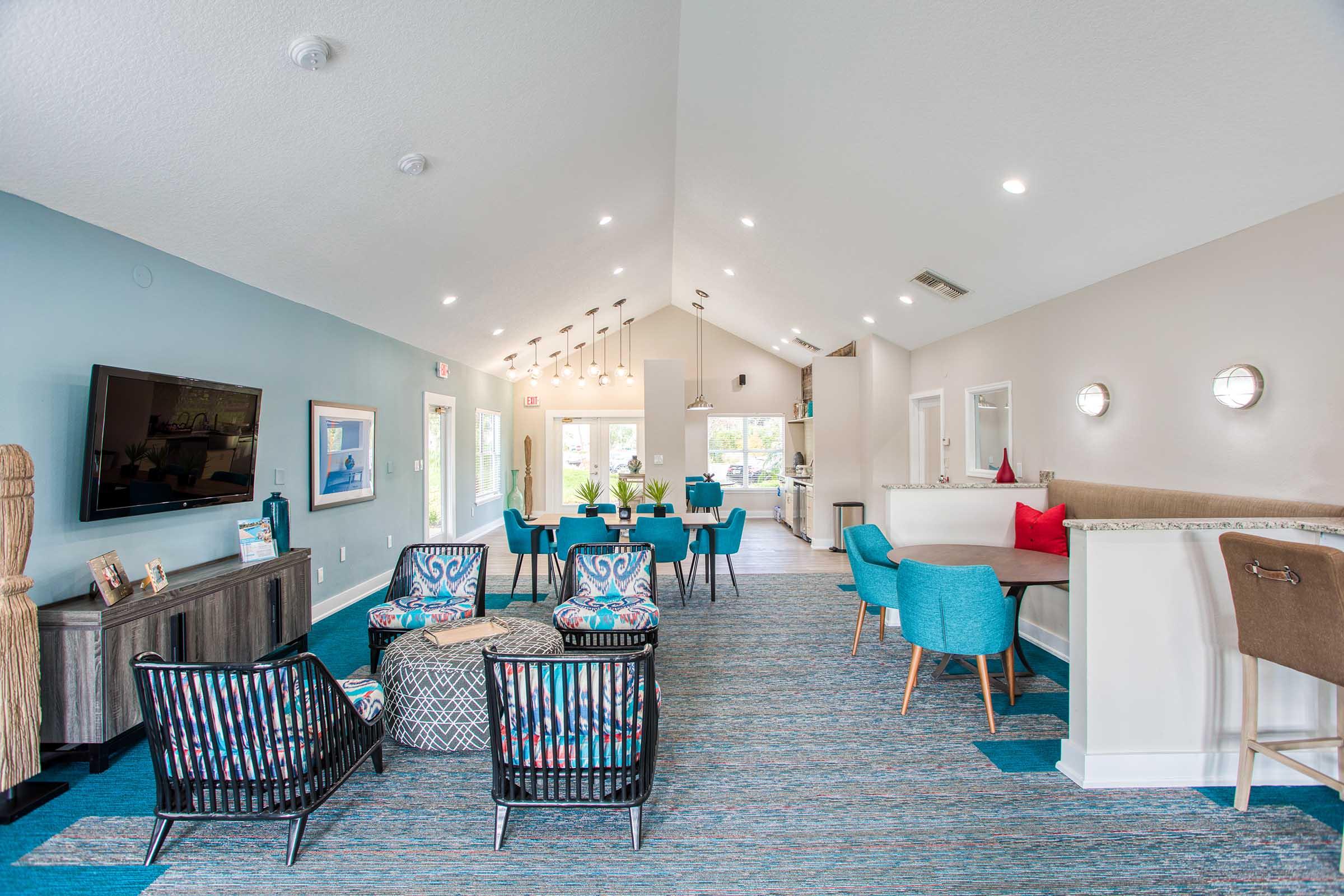 A modern and inviting lounge area featuring blue and patterned chairs, a large flat-screen TV on the wall, and natural light streaming through windows. There are decorative plants and cozy seating arrangements, creating a welcoming atmosphere for relaxation or socializing.