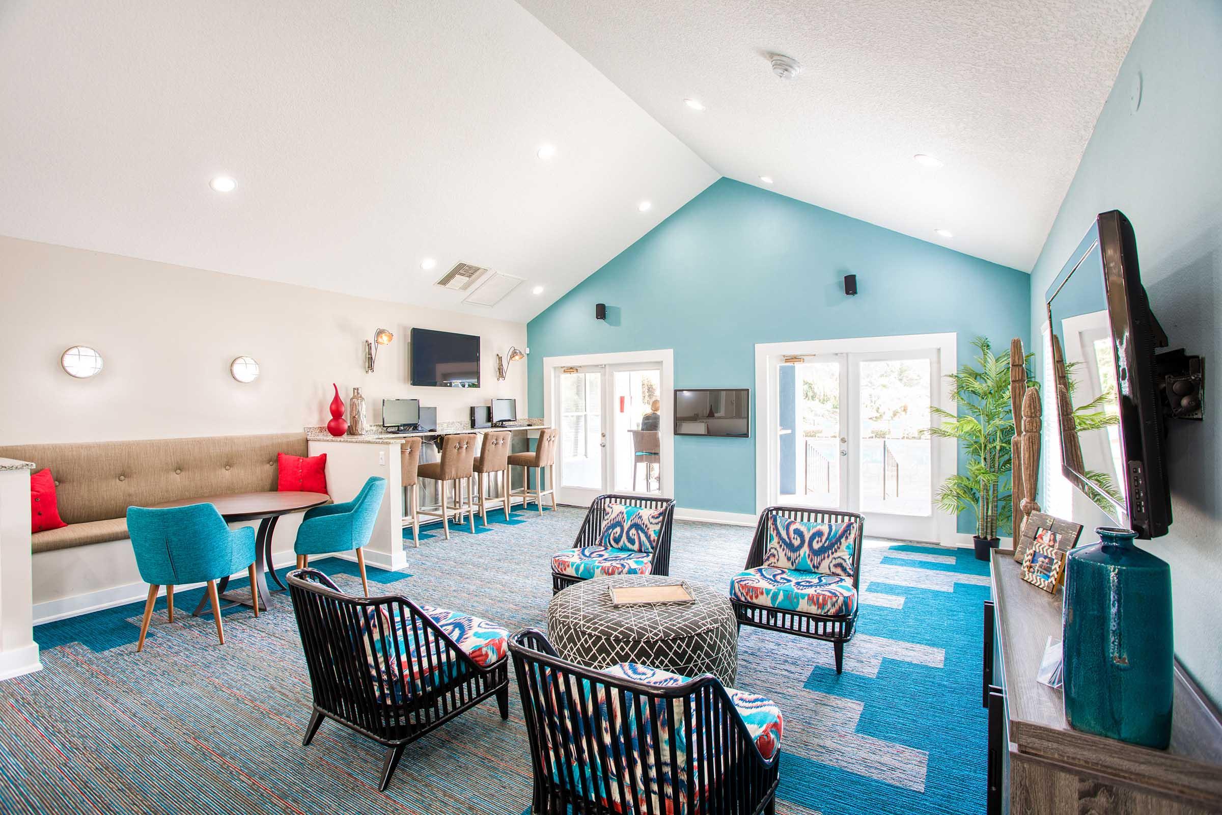 Interior of a bright and modern lounge area featuring a high ceiling, colorful seating, and large windows. The decor includes a mix of casual furniture, such as patterned chairs and a cozy couch, with a vibrant rug and wall accents in shades of blue and white. A flat-screen TV is mounted on the wall.