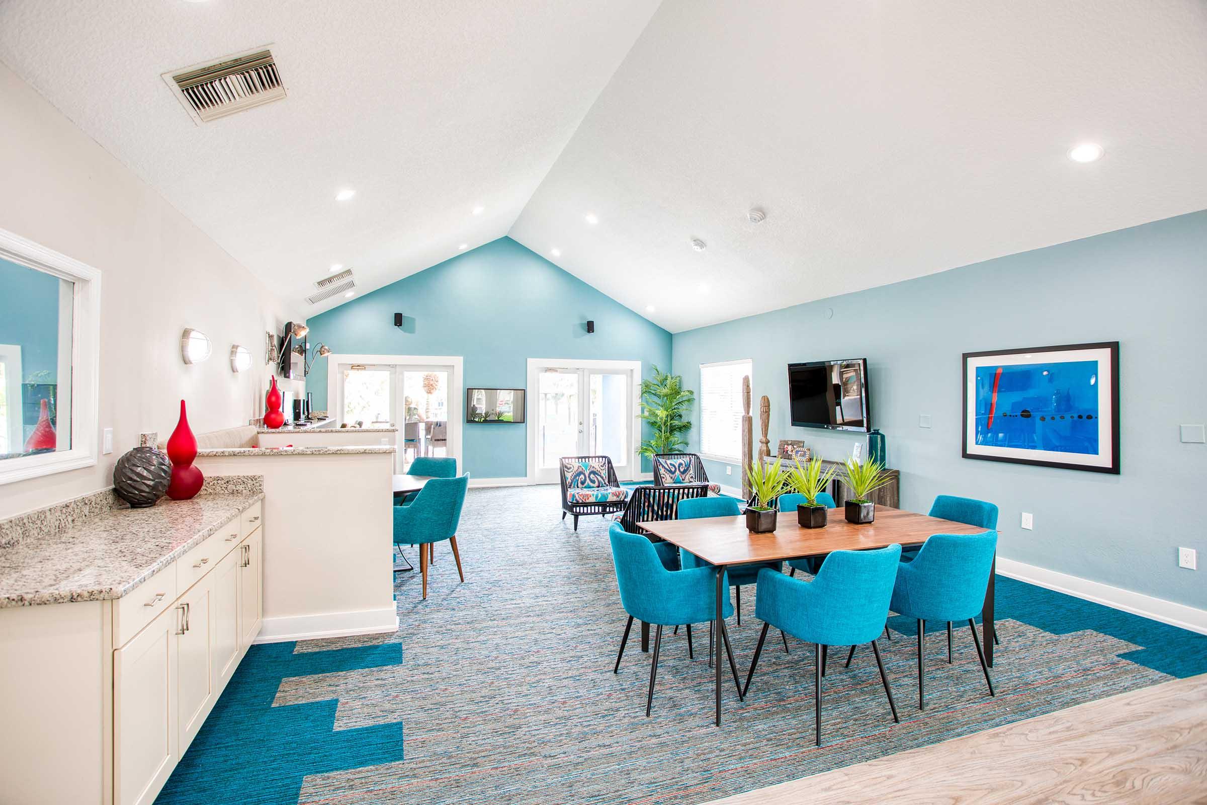 A bright and modern community room featuring a dining area with a large wooden table surrounded by turquoise chairs. The room has a high ceiling, light gray walls, and blue carpeting. Large windows allow natural light, and decorative plants and artwork add style to the space.