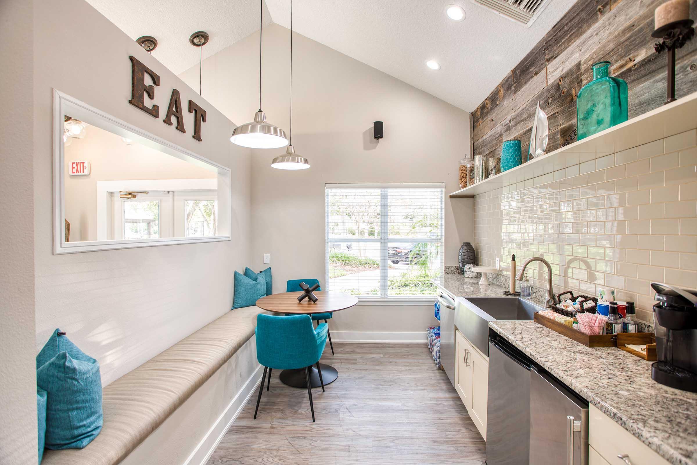 A bright and modern kitchen area with a cozy dining nook featuring a round table and teal chairs. A bench with pillows lines the wall, and there are decorative items on shelves. The space has large windows allowing natural light and a warm color scheme, creating an inviting atmosphere for dining.