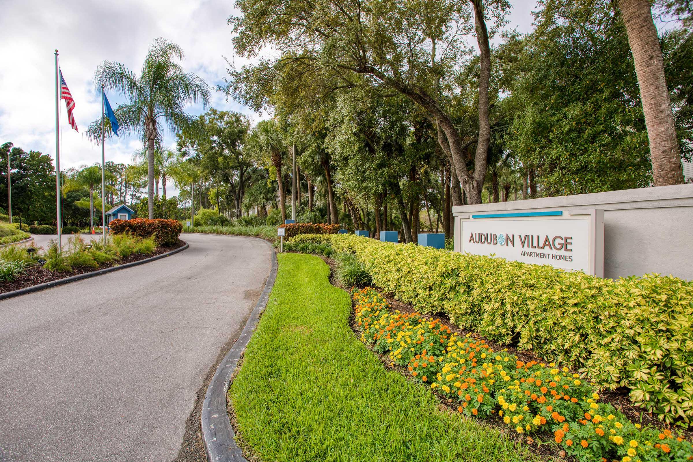 Entrance to Audubon Village Apartment Homes, featuring a curved driveway lined with lush greenery and colorful flower beds. American flags are visible in the background, along with palm trees and other landscaping elements, creating a welcoming atmosphere at the entrance.