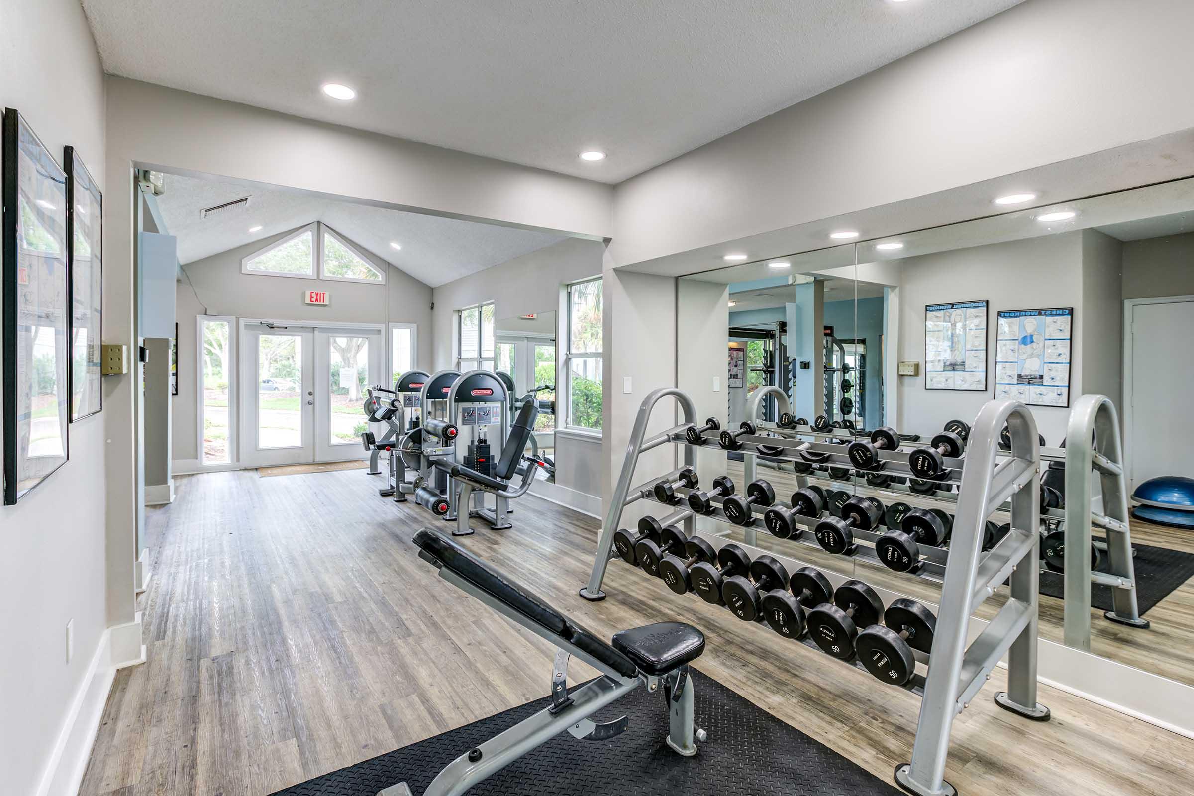 A modern fitness center featuring cardio machines, a weight bench, a rack of dumbbells, and large mirrors. The space is bright with natural light coming through large windows and is designed for a comfortable workout experience.