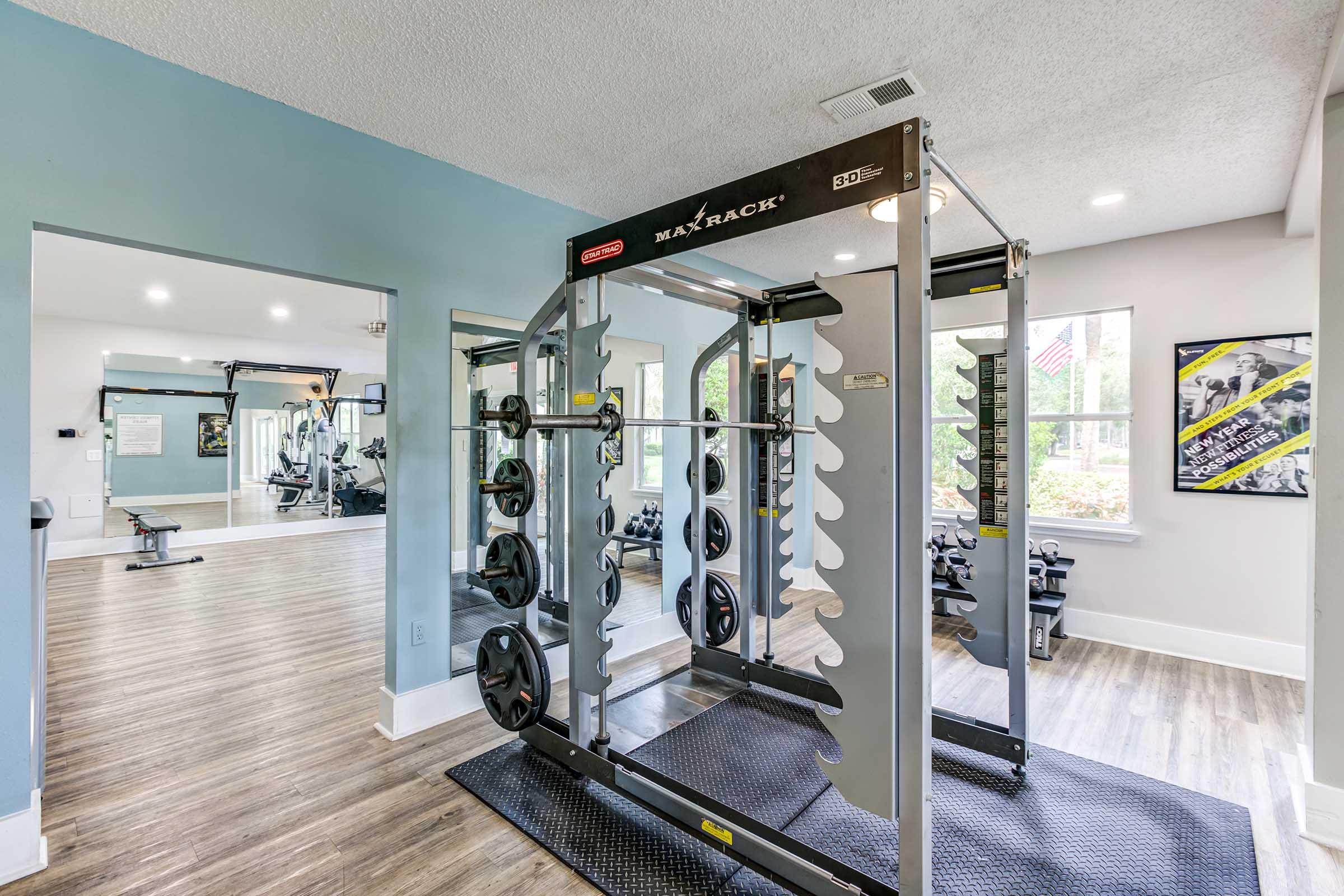 A well-equipped gym featuring a squat rack with weights, set against a backdrop of large windows letting in natural light. The floor is wooden, and the space includes various fitness machines and exercise equipment, creating a bright and inviting workout environment.
