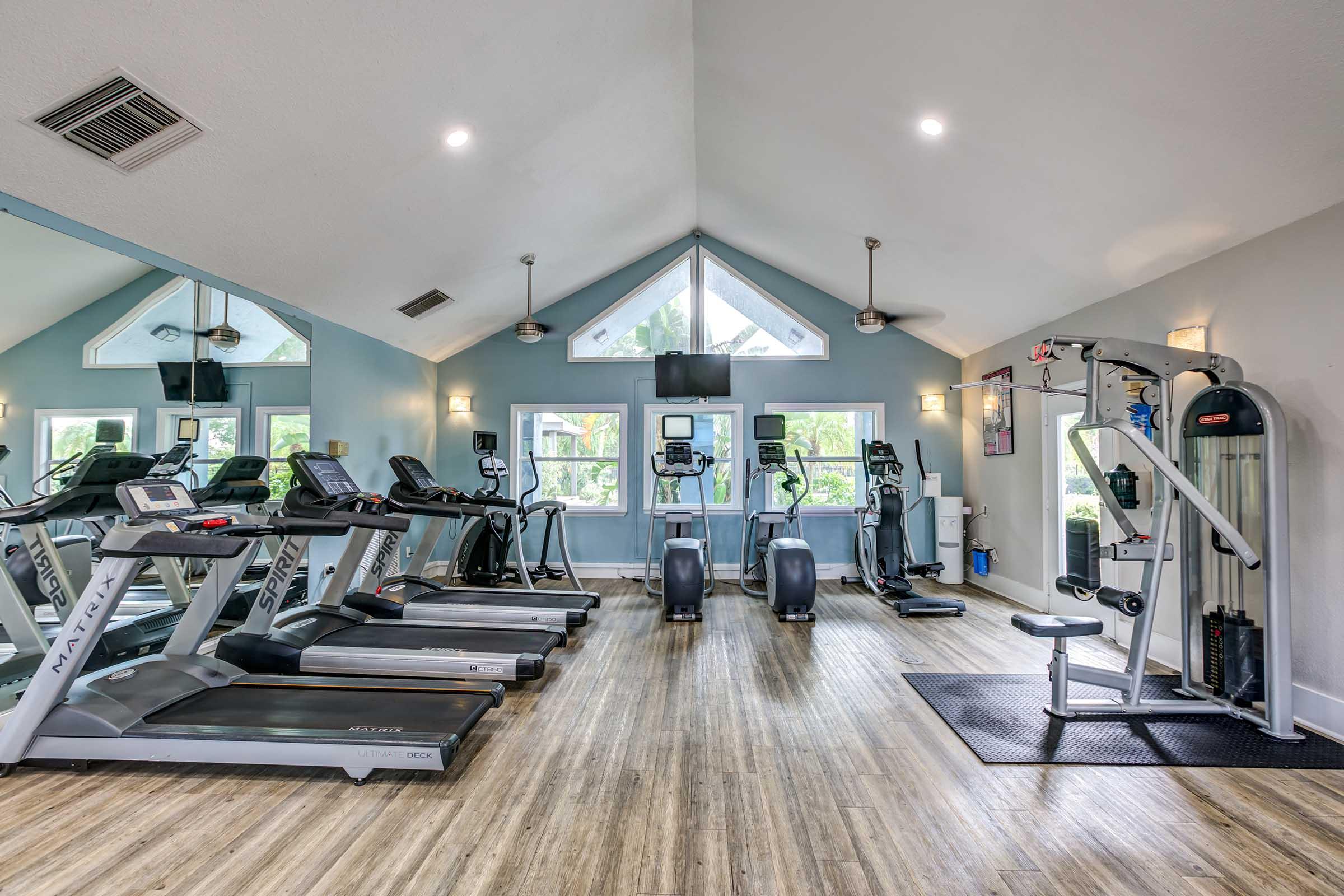 A bright and modern gym with large windows, featuring several treadmills and elliptical machines lined against the walls, a multi-station weight machine, and a black exercise mat. The wooden floor adds warmth to the space, while overhead lights provide ample illumination.