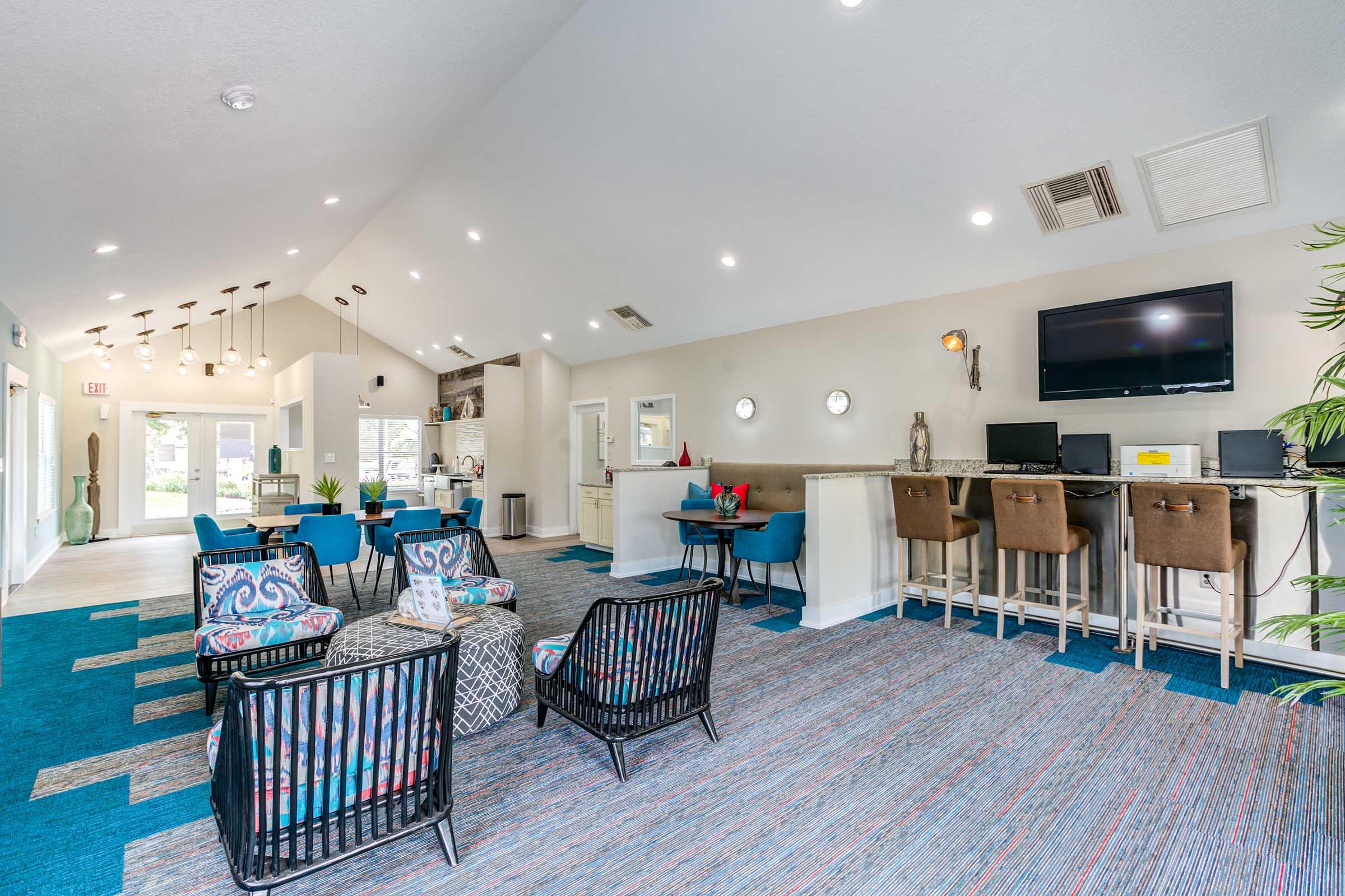 A modern lounge area featuring comfortable seating with colorful chairs and tables. The space includes a bar with high stools, a large television, and decorative plants. Bright lighting enhances the inviting atmosphere, with a view of the entrance and kitchen area in the background.