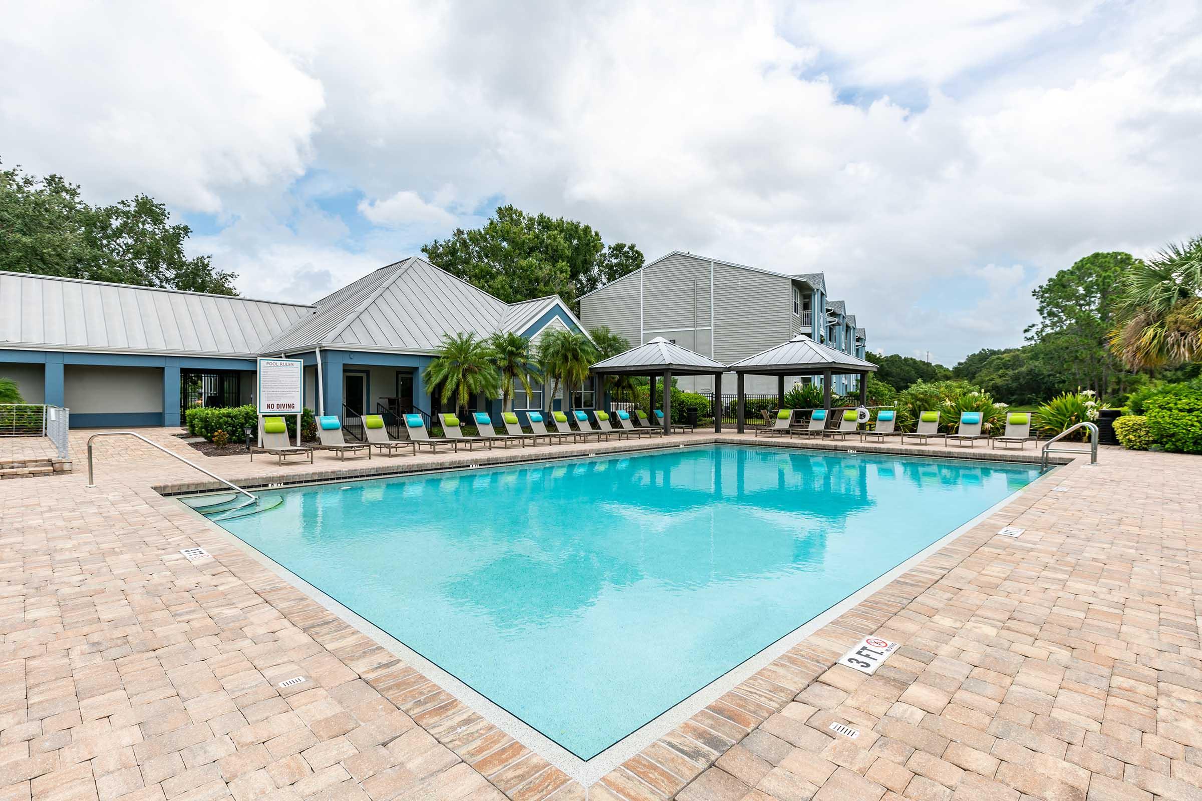 A serene swimming pool surrounded by lounge chairs, palm trees, and a gazebo. The area features a brick patio and is set against a backdrop of lush greenery and residential buildings under a cloudy sky. Perfect for relaxation or leisure activities.