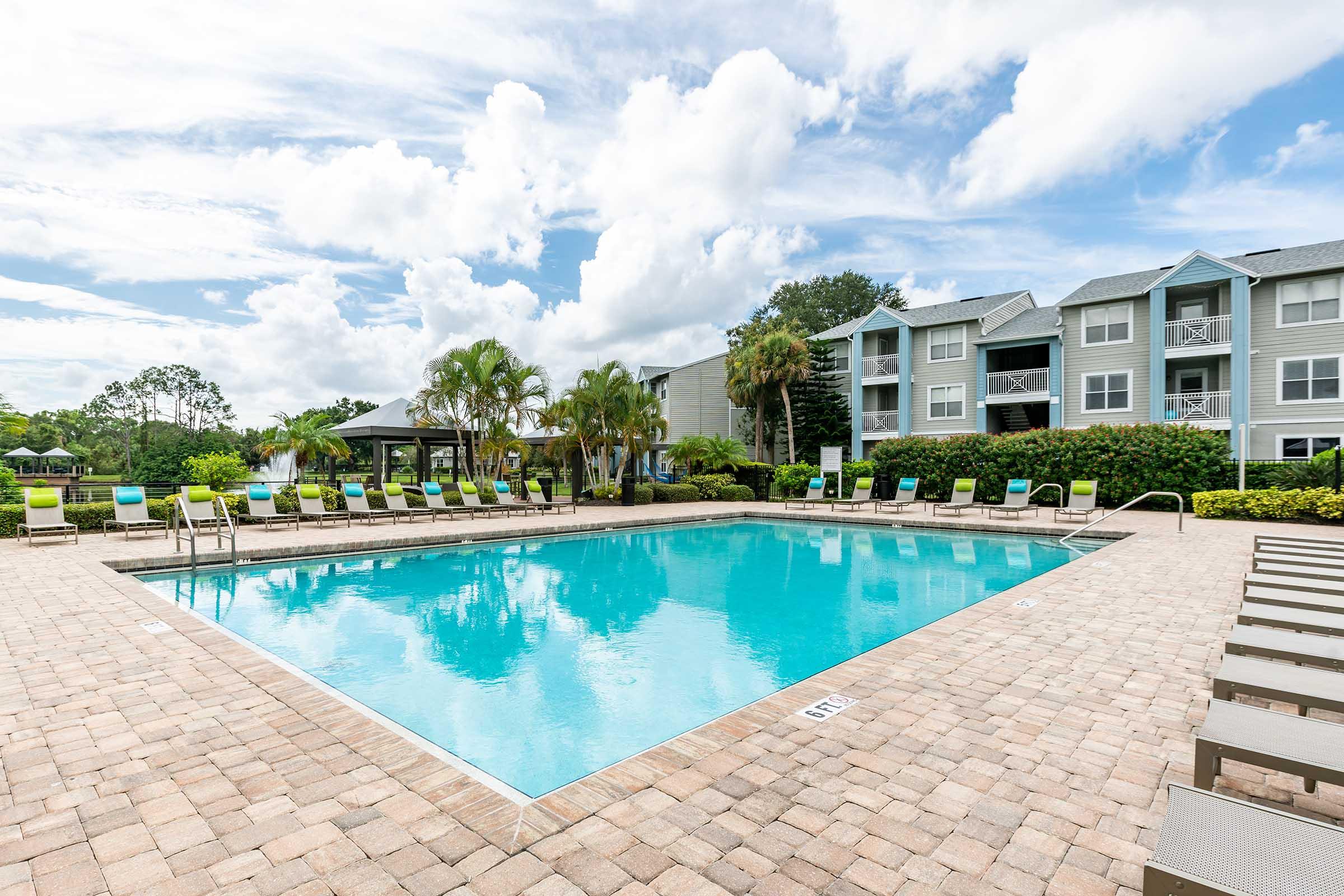 A large outdoor swimming pool surrounded by lounge chairs, set against a backdrop of green palm trees and a blue sky with fluffy clouds. In the background, there are multi-story apartment buildings with balconies overlooking the pool area.