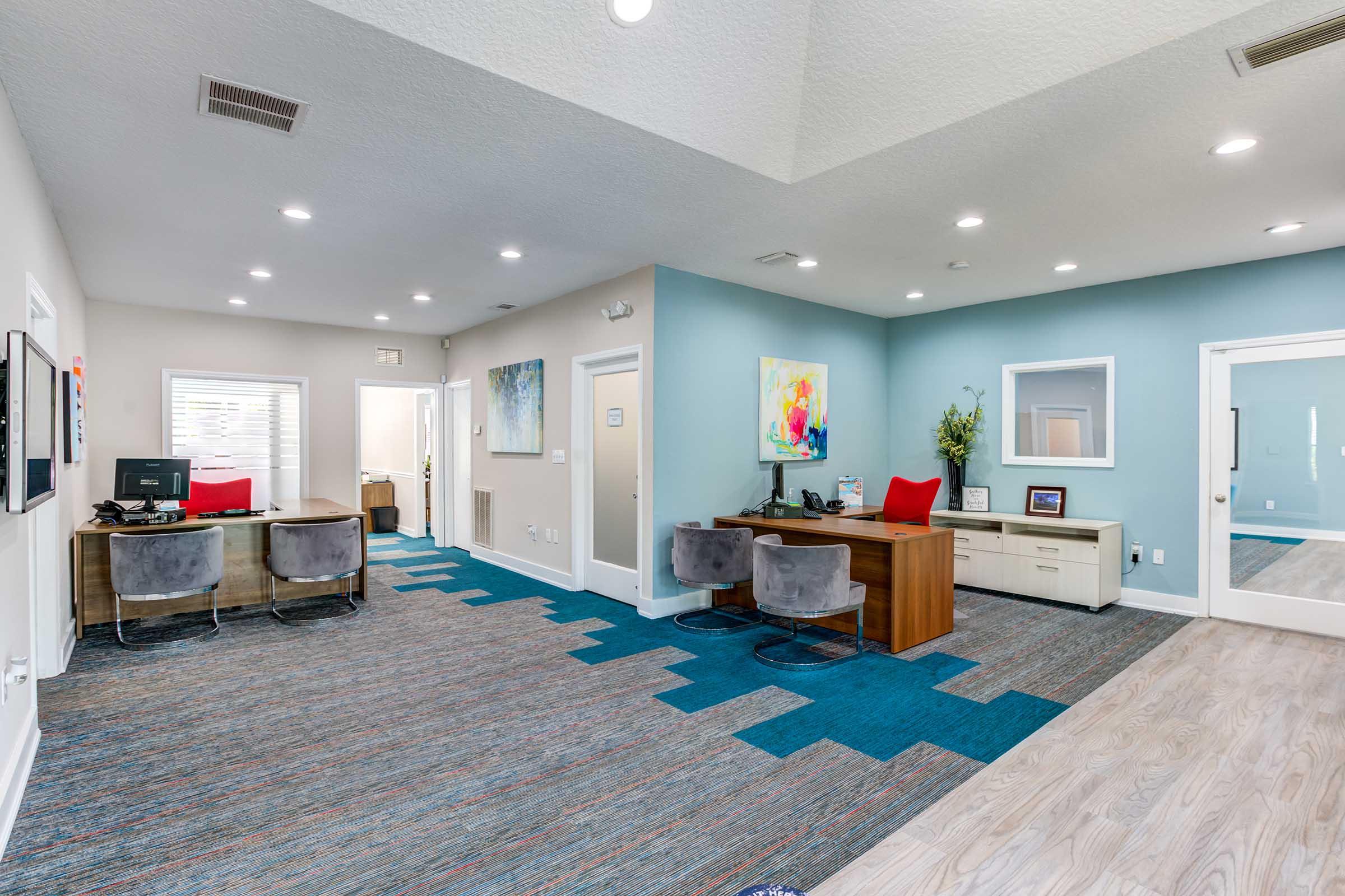 A modern office space featuring light gray walls and a blue accent wall. It includes a workstation with a desktop computer, gray chairs, and a desk with a red accent chair. The flooring combines light wood and vibrant blue carpet tiles. Natural light streams through windows, creating a welcoming atmosphere.
