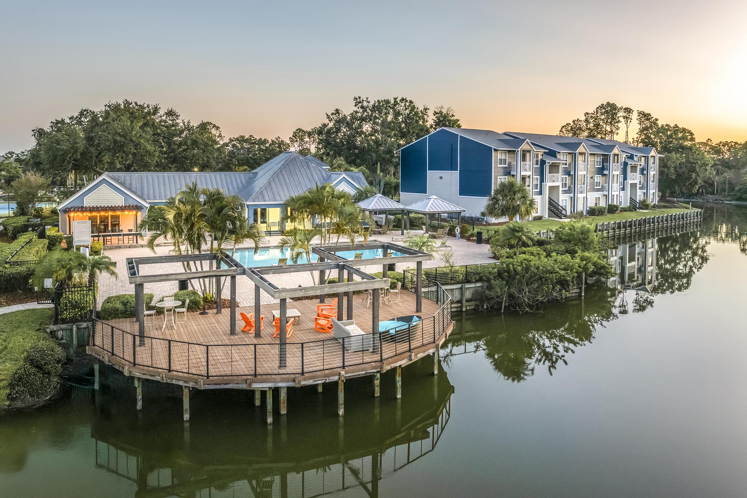 A scenic view of a waterfront property featuring a swimming pool, palm trees, and a wooden deck with orange chairs. The background includes modern apartment buildings and lush greenery, with calm water reflecting the colorful sky at sunset.