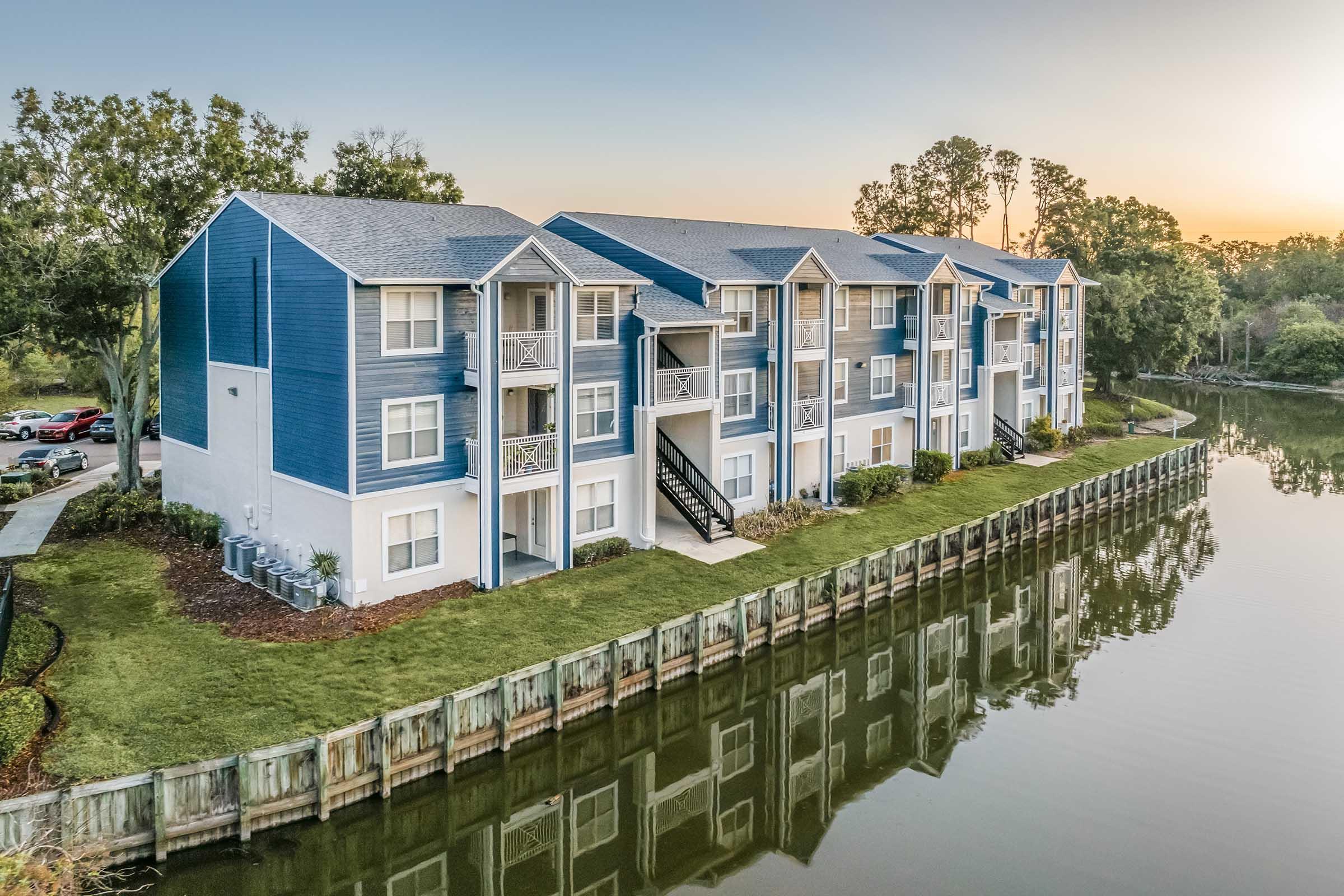 A modern apartment building with blue and white exterior, situated by a calm lake. The structure features multiple balconies, lush green grass in front, and a wooden dock along the water's edge. The scene is illuminated by a soft sunset, reflecting on the lake's surface.