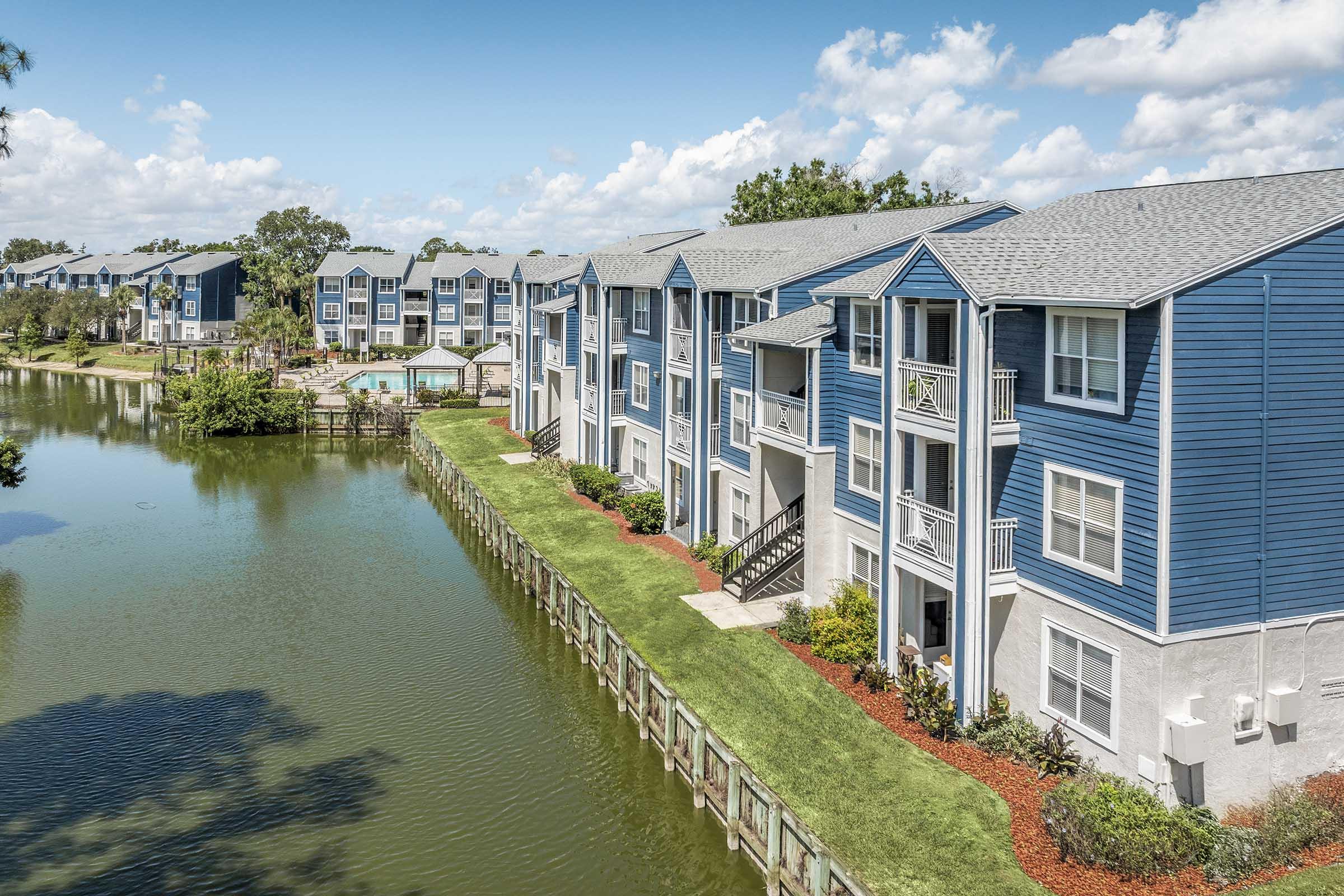 A vibrant residential complex featuring blue and gray apartment buildings along a scenic lake. The landscape includes well-maintained lawns, lush greenery, and a pool area visible in the distance under a bright blue sky with fluffy white clouds.