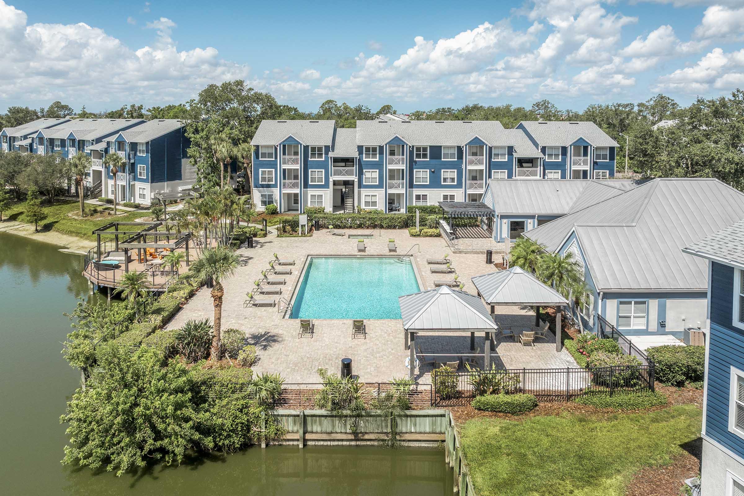 Aerial view of an apartment complex featuring a large swimming pool surrounded by lounge chairs. Lush greenery and palm trees are visible around the pool area, with several buildings in the background. The sky is bright blue with fluffy white clouds.