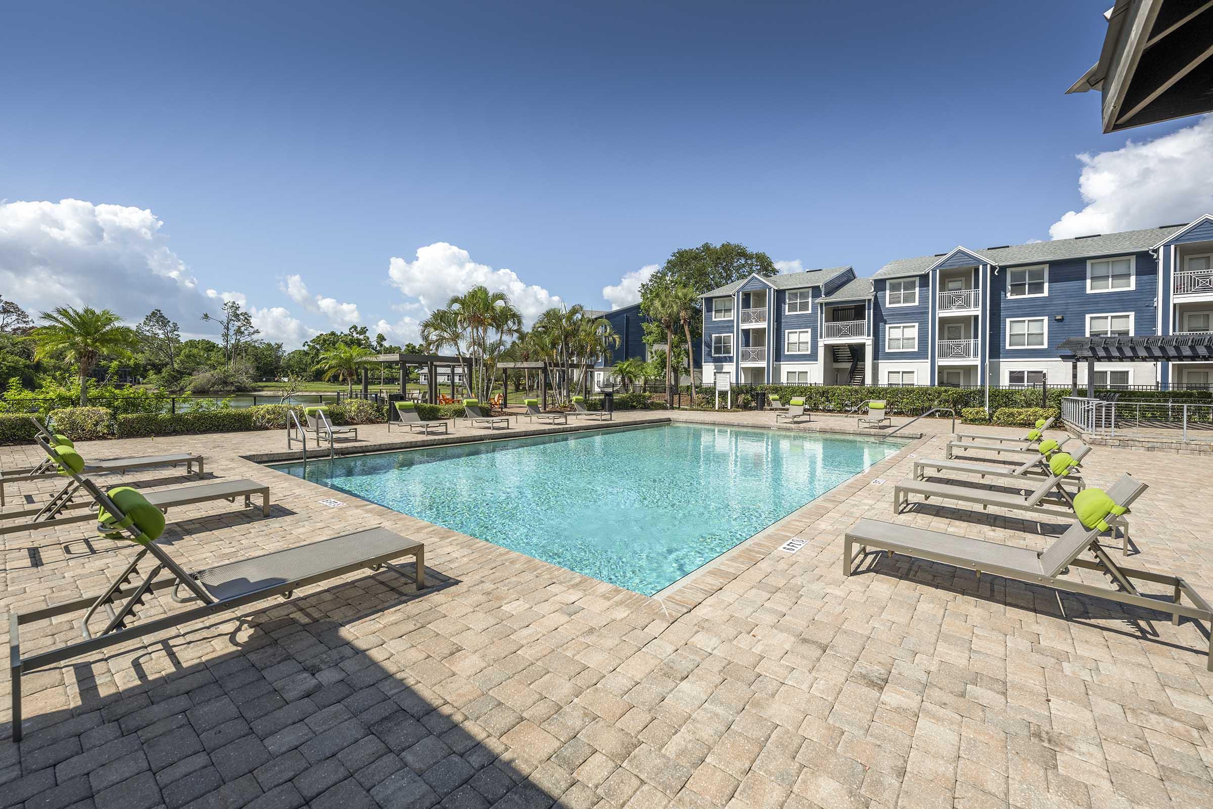 A clear blue swimming pool surrounded by lounge chairs on a brick patio, set against a backdrop of green landscaping and a multi-story building. The sky is bright with a few white clouds, creating a sunny atmosphere perfect for relaxation.