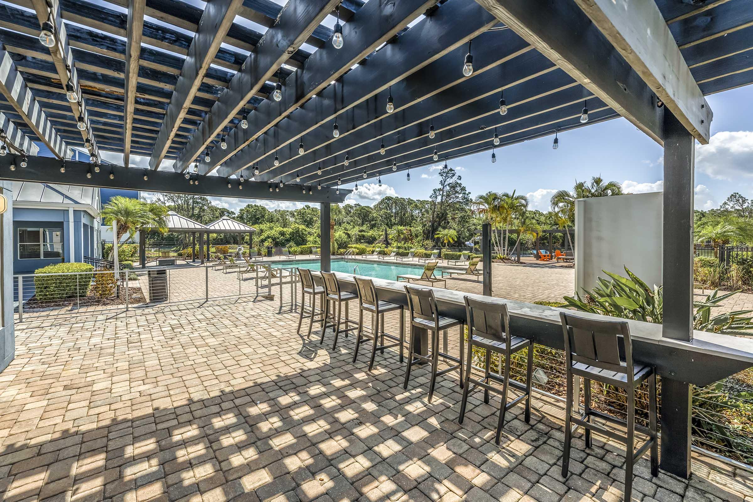 A spacious outdoor area with a pergola featuring string lights, set beside a swimming pool. There are several bar stools lined up along the structure, and lush greenery and lounge chairs are visible in the background. The scene is bright, showcasing a relaxing atmosphere.