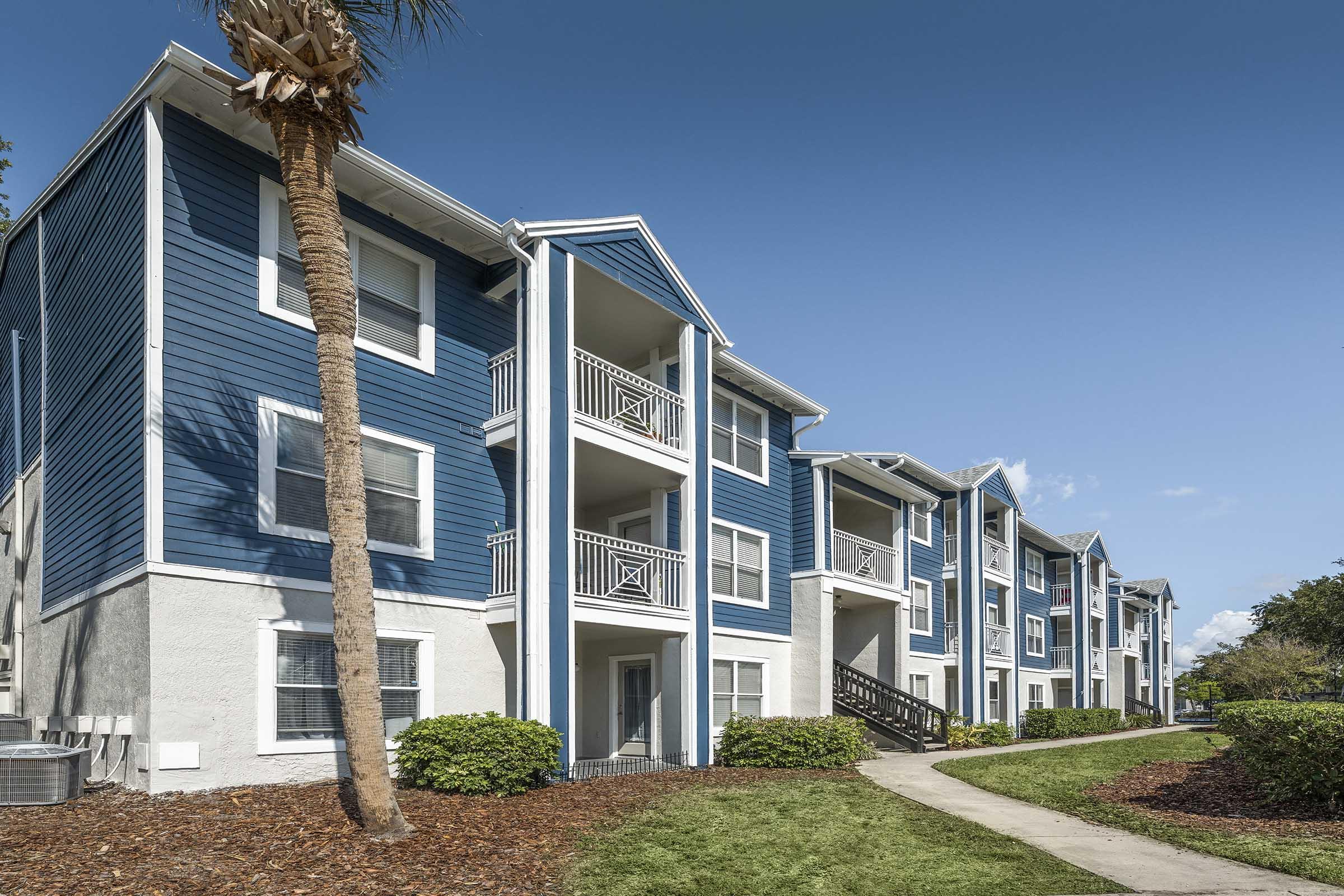 A row of modern apartment buildings with blue siding and white accents, featuring balconies and landscaped greenery. A palm tree is visible in the foreground, and a clear blue sky is in the background, creating a vibrant atmosphere. The walkway leads to the entrance of the buildings.