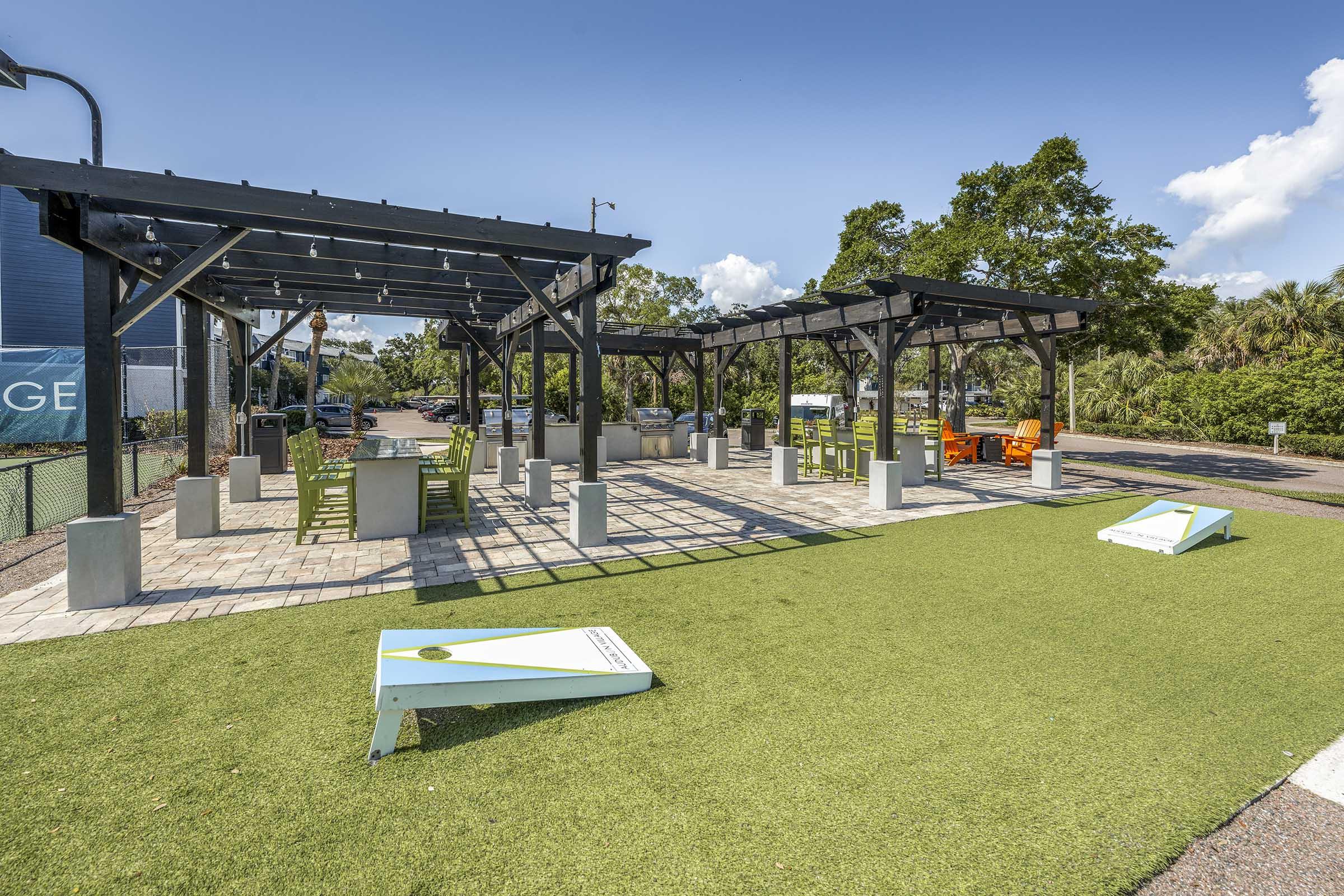A well-lit outdoor recreational area featuring covered seating structures, green artificial turf, and cornhole boards. The space is surrounded by trees and has a clear blue sky with scattered white clouds, providing a pleasant environment for social activities and games.