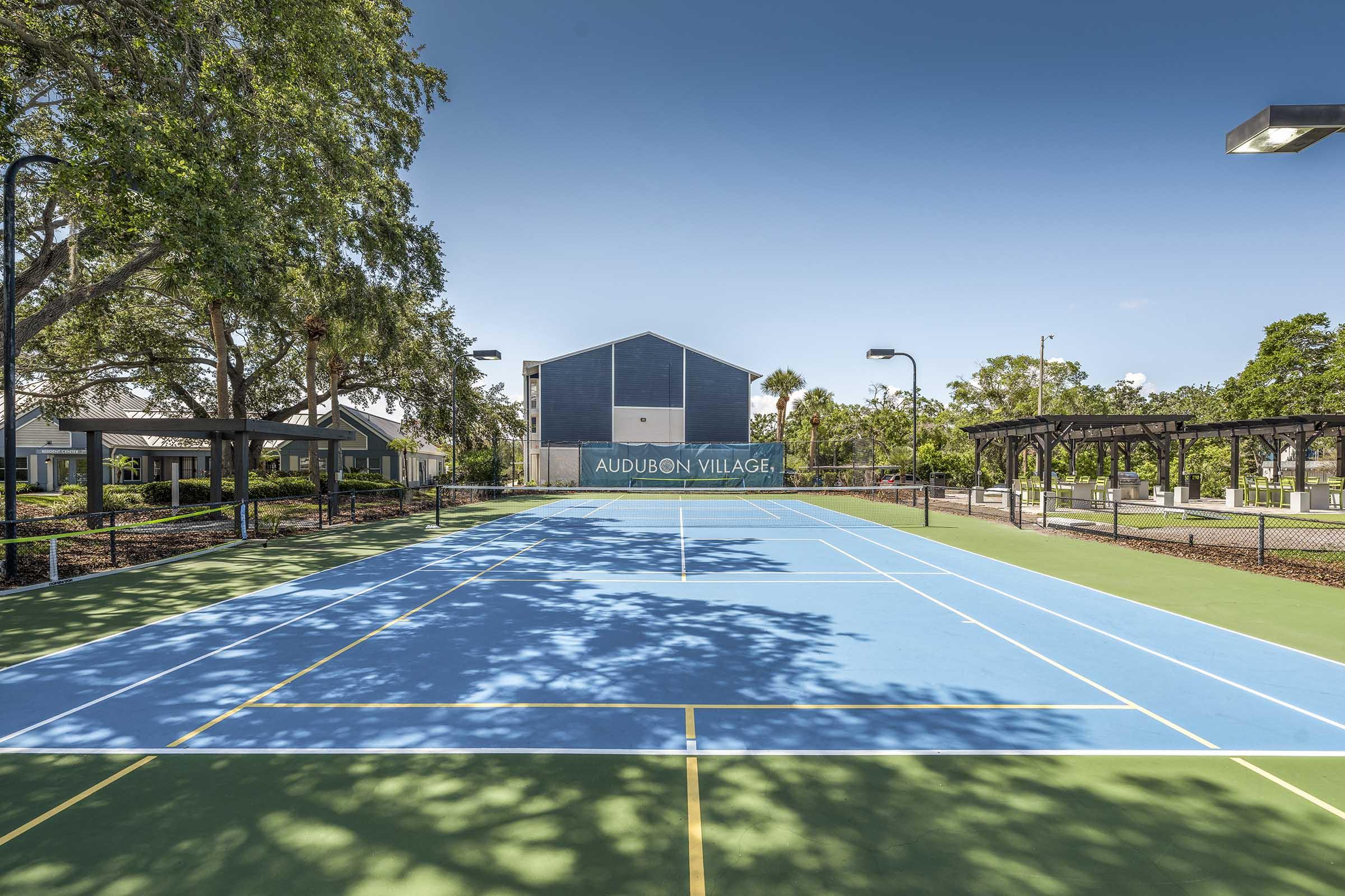 A bright, sunny tennis court with a blue playing surface and green sidelines, set against a backdrop of trees. In the distance, a modern building with the text "AUDUBON VILLAGE" is visible. The scene captures a clear sky and well-maintained outdoor recreational area.