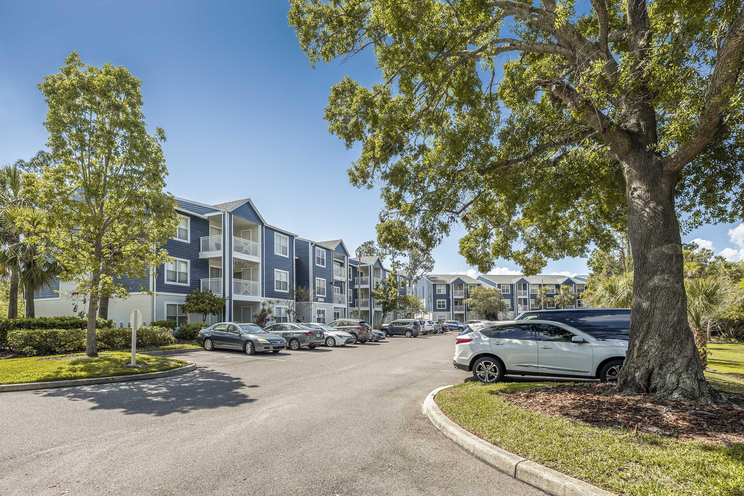 A residential apartment complex surrounded by lush greenery and trees. The street is lined with parked cars, and the buildings feature a modern design with blue and white exteriors. Bright blue skies and scattered clouds are visible above, indicating a sunny day.
