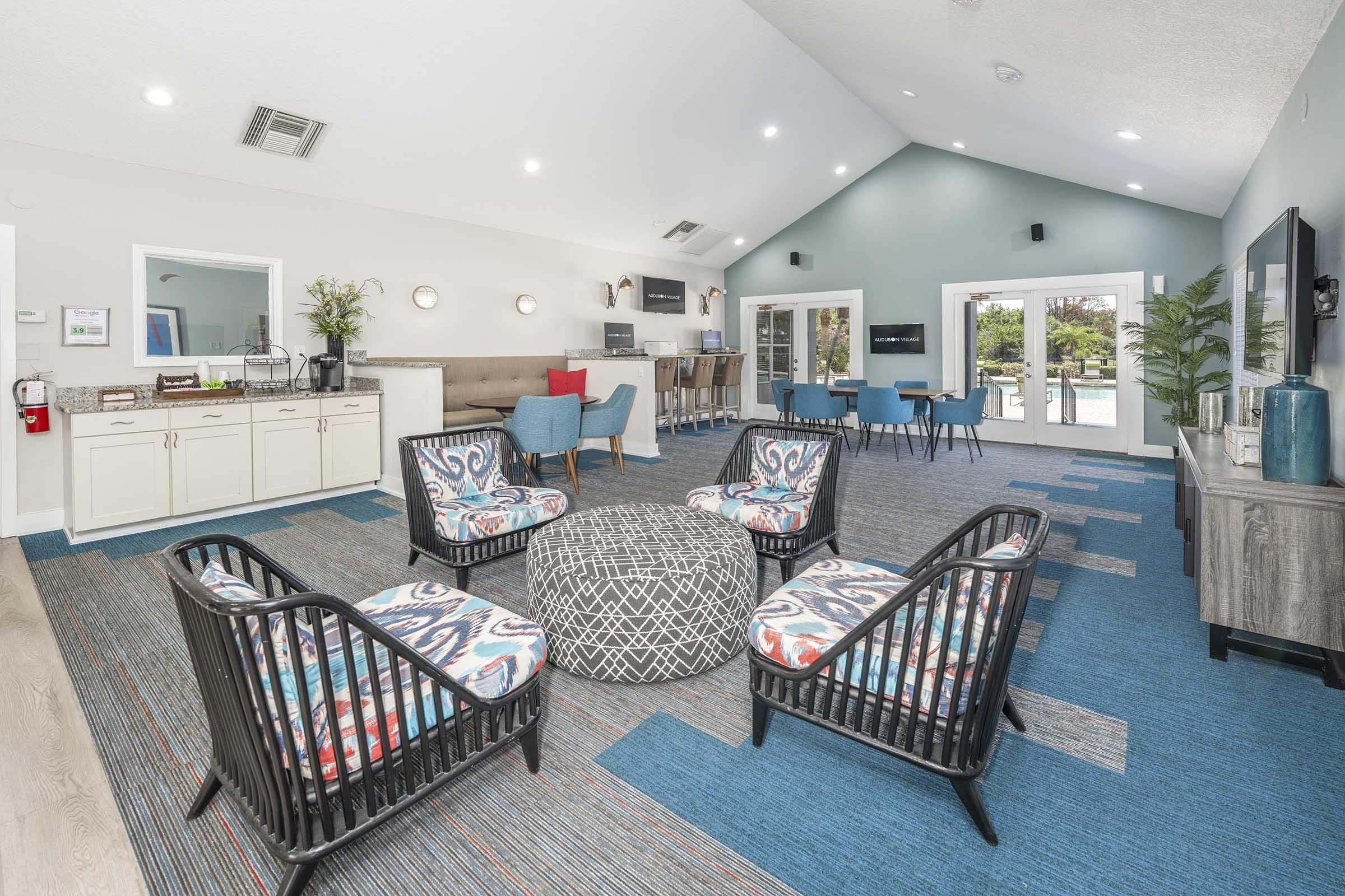 Interior of a modern lounge area featuring several black wicker chairs with colorful cushions arranged around a patterned ottoman. There are barstools along a counter, a large window with views outside, and blue and grey decor on the walls. A reception area with plants is also visible.