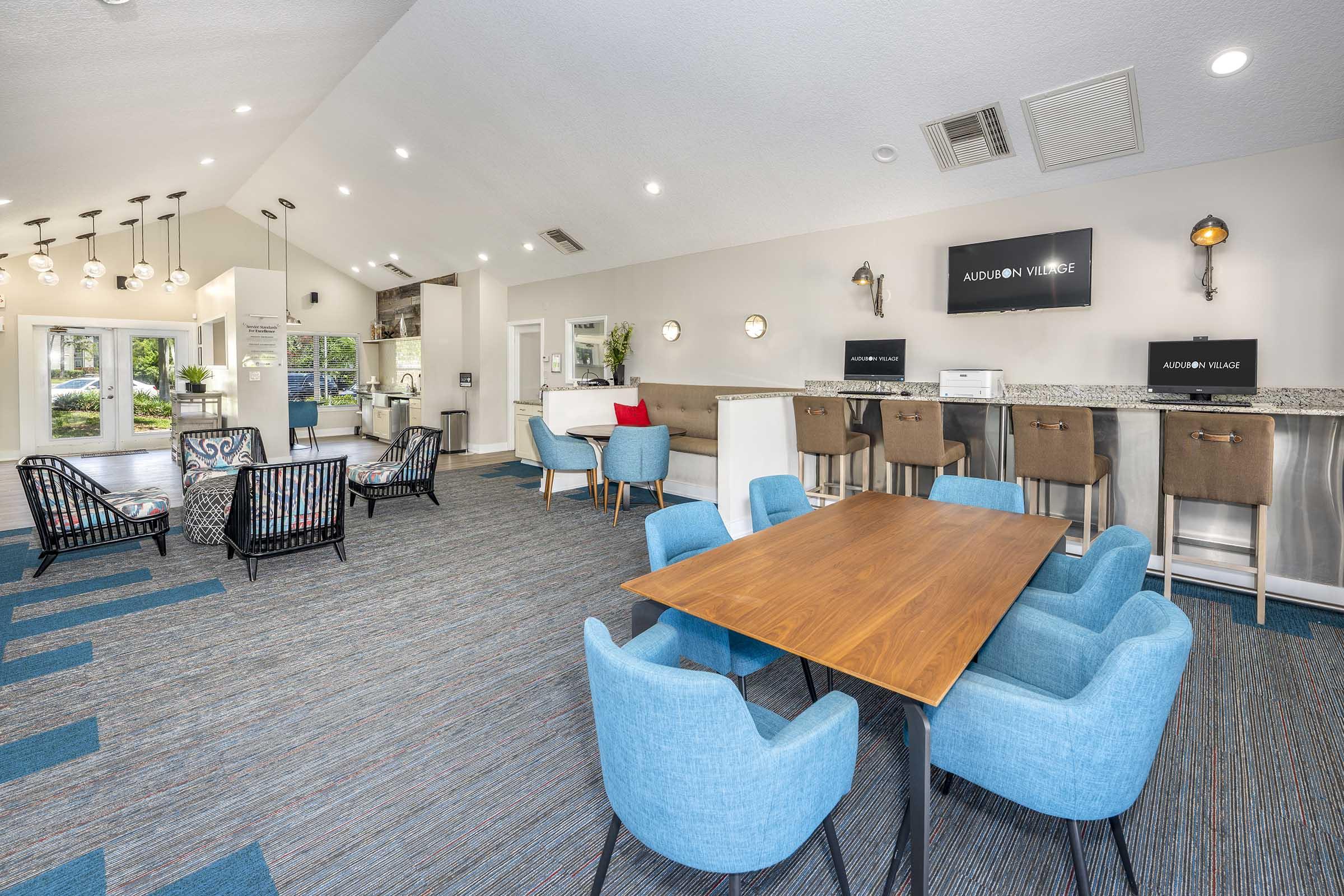 A modern, spacious communal area featuring a large wooden table surrounded by blue chairs, with two stylish armchairs nearby. There is a bar area with high stools and a kitchenette visible in the background. The decor includes contemporary lighting and a welcoming ambiance, perfect for social interactions.
