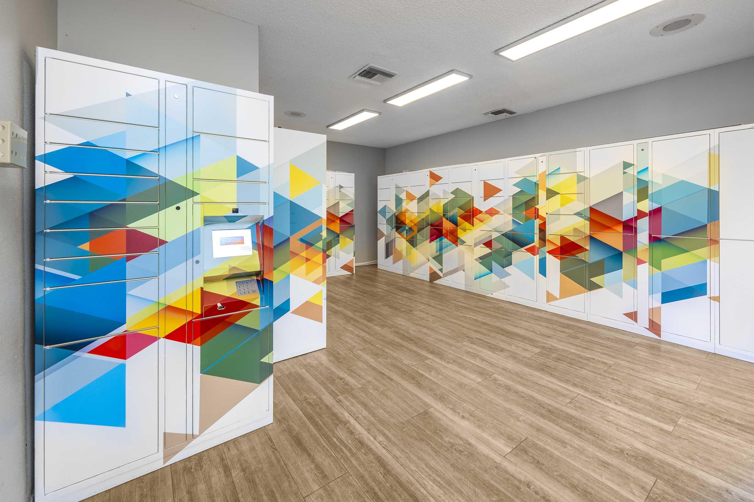 A modern locker room with bright, geometric murals on the lockers. The space features multiple lockers with a variety of colorful designs in blues, greens, and oranges on a neutral wall. The floor is wooden, and there is good overhead lighting illuminating the area.