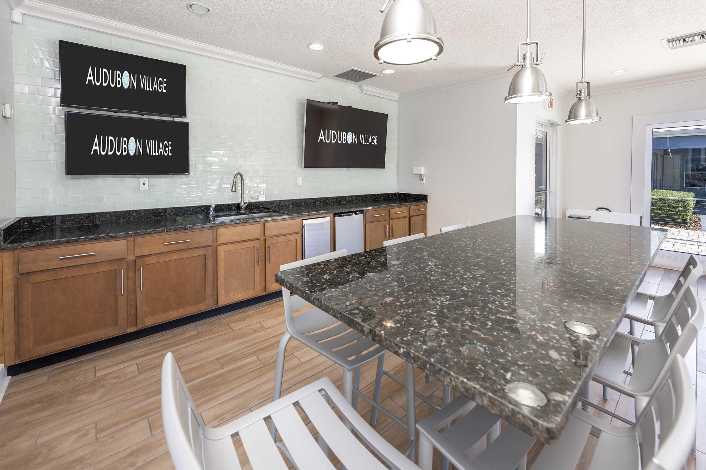 Modern kitchen space featuring a large granite island with seating, wooden cabinetry, and stainless steel appliances. Two wall-mounted screens display the text "AUDUBON VILLAGE." The bright, airy room has a light color scheme with natural wood flooring and ample lighting.