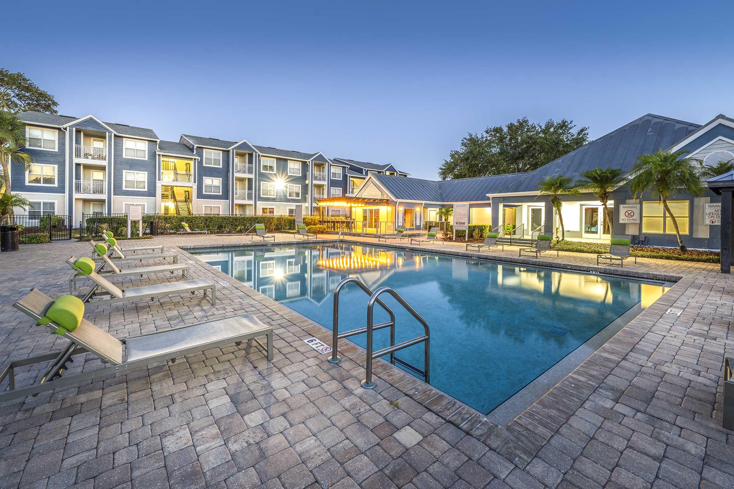 A serene swimming pool surrounded by lounge chairs, framed by trees and modern apartment buildings. The pool reflects the evening sky, and there are illuminated areas near the poolside. The setting conveys a peaceful and inviting atmosphere for relaxation and leisure.