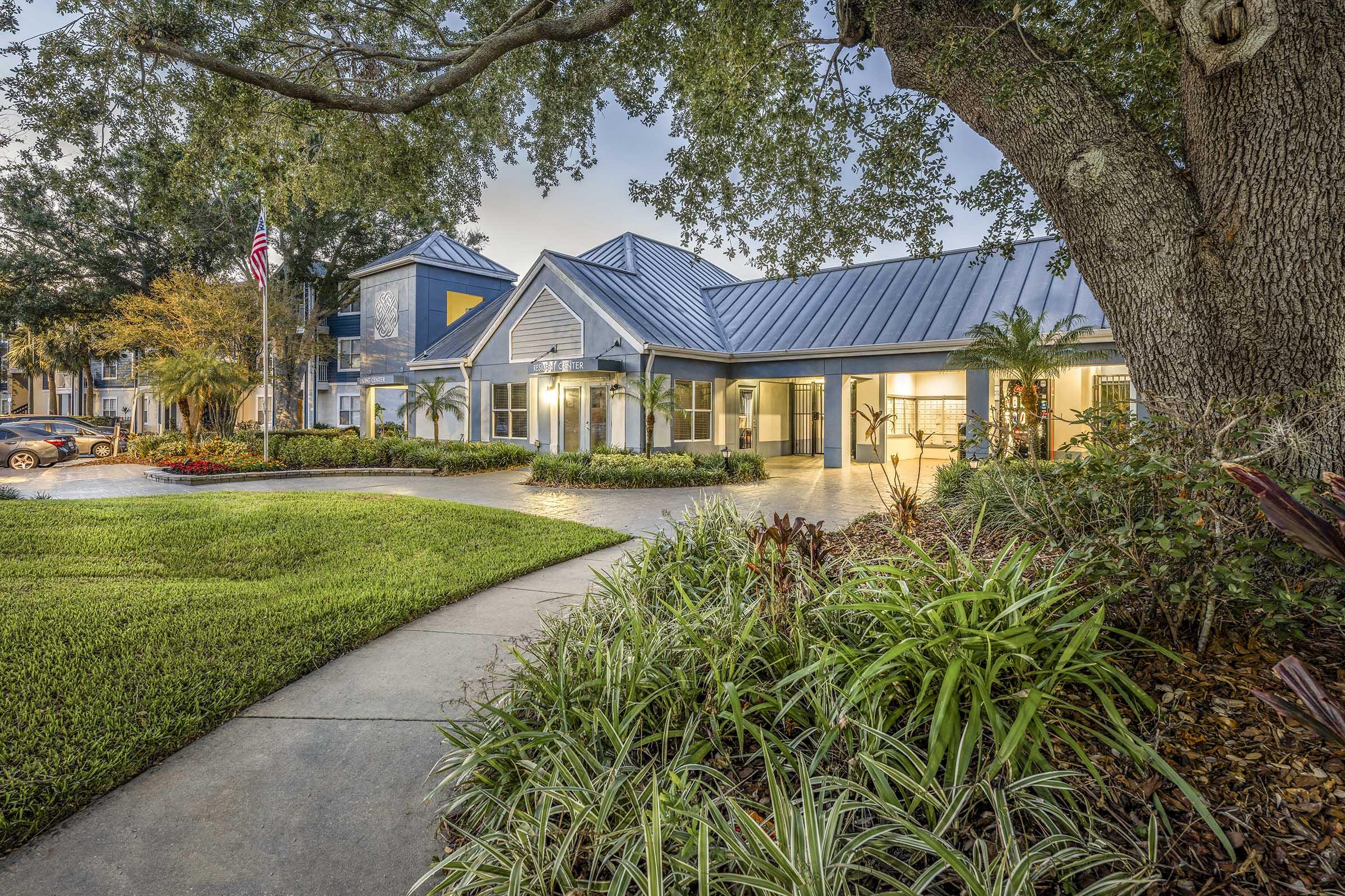 A modern building with a blue and gray exterior, featuring a sloped metal roof, surrounded by lush green landscaping and palm trees. A well-maintained walkway leads to the entrance, with American flags and flowers accentuating the scene, set during twilight with soft lighting.