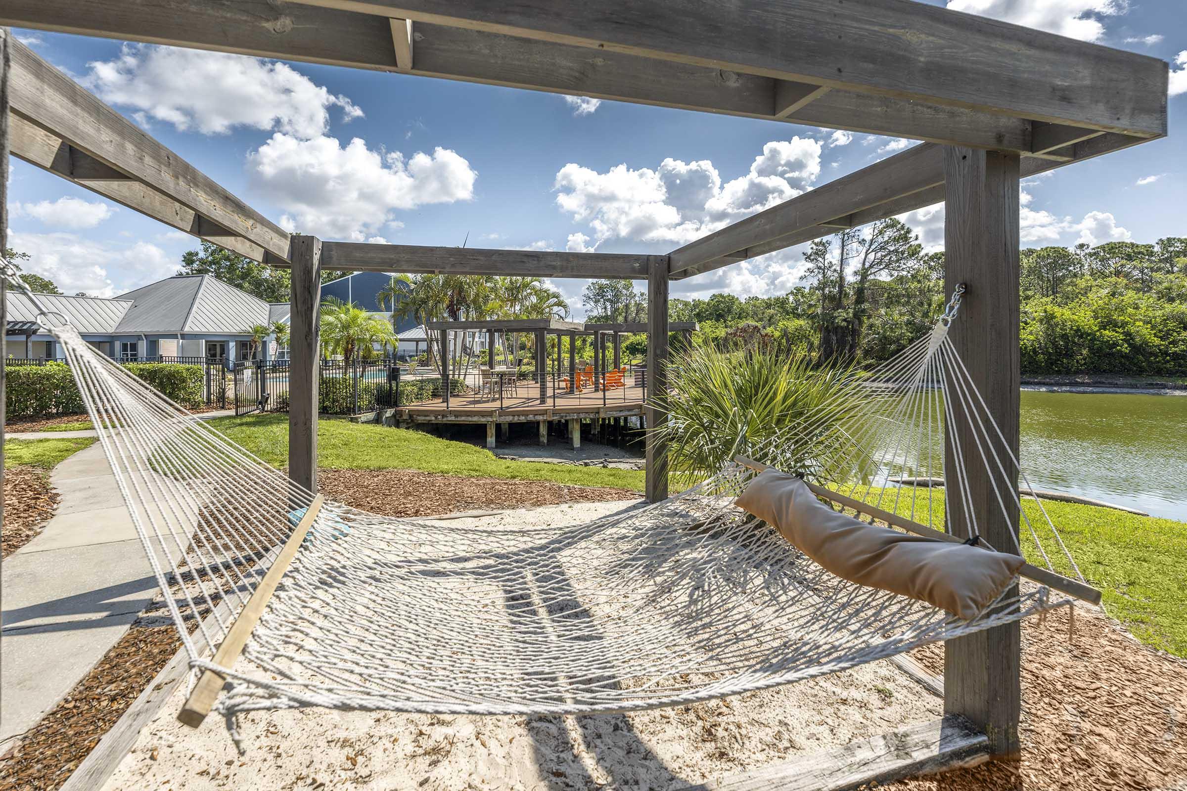 A tranquil outdoor scene featuring a hammock suspended between wooden supports, set against a backdrop of a serene lake, lush greenery, and a blue sky with fluffy clouds. In the distance, there are picnic areas and wooden decking, creating a peaceful and inviting atmosphere.
