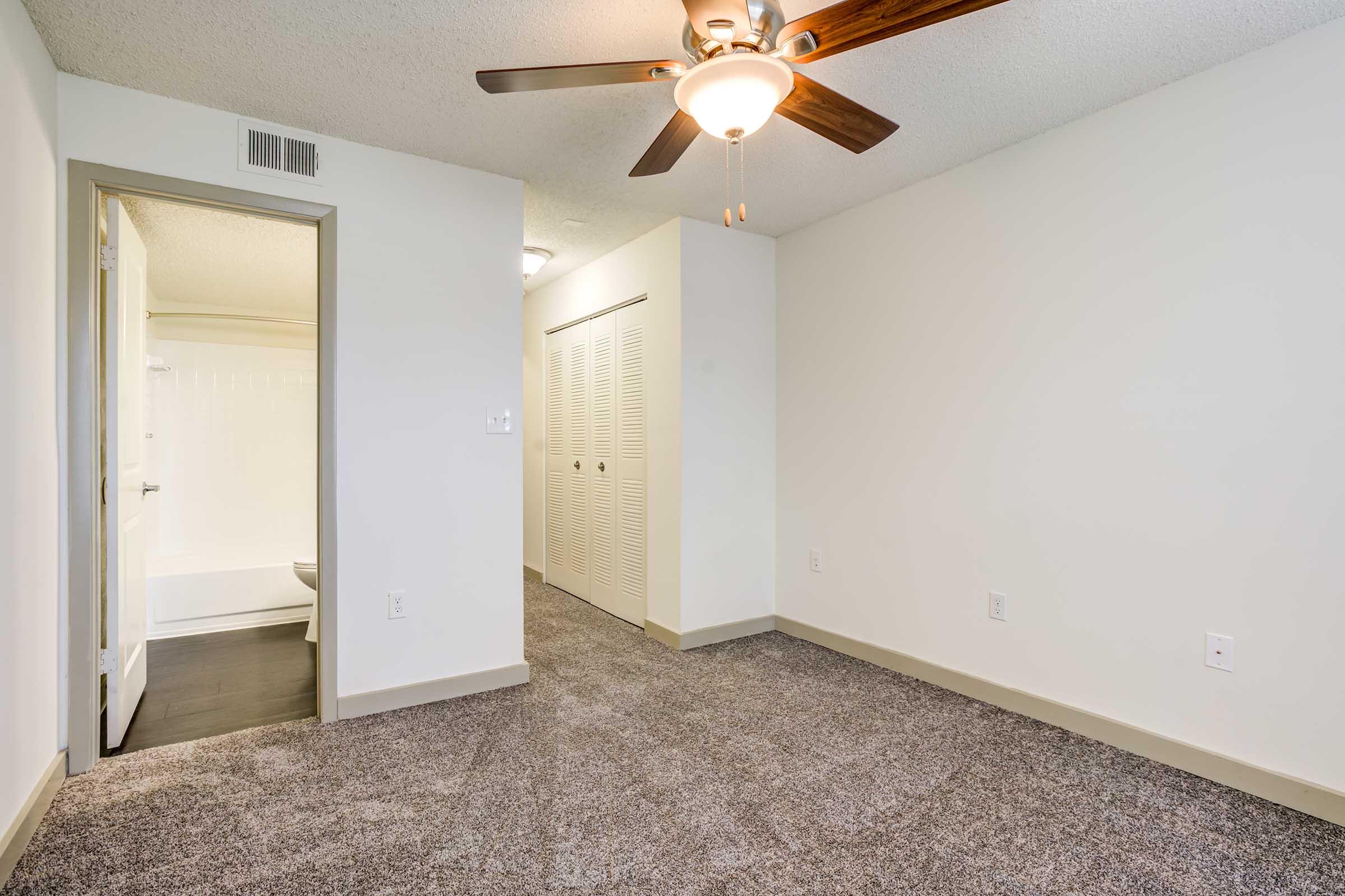 A well-lit room featuring a ceiling fan, neutral-colored walls, and plush carpet. To the left, there is an open door leading to a bathroom, while the right side has a closet with sliding doors. The layout is spacious and inviting, with soft natural light enhancing the atmosphere.