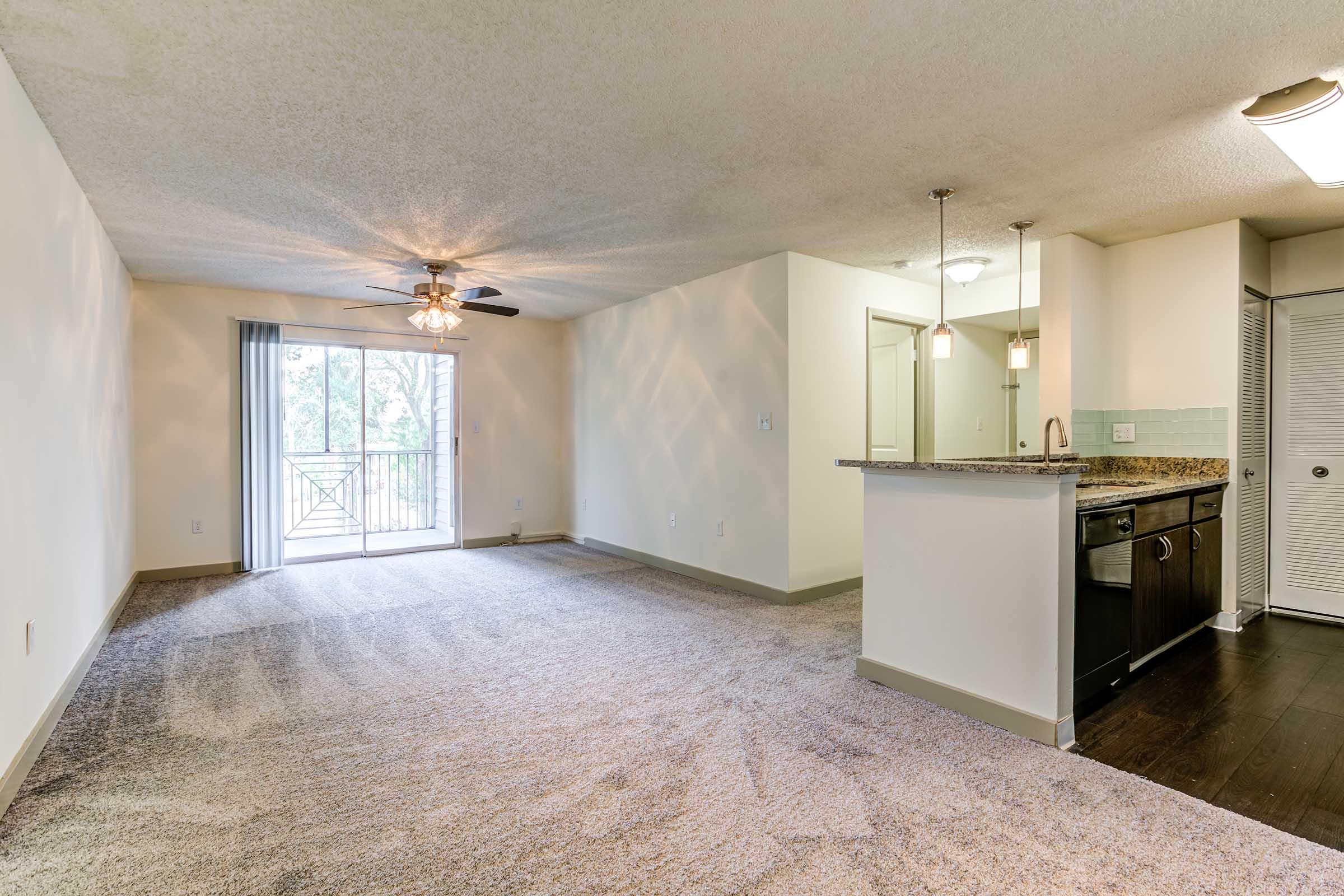 Spacious living area featuring plush carpet, a ceiling fan, and sliding glass doors leading to a balcony. Modern kitchen with granite countertops, dark cabinetry, and stainless steel appliances. Natural light filters in, enhancing the open layout. Neutral colors create a warm, inviting atmosphere.