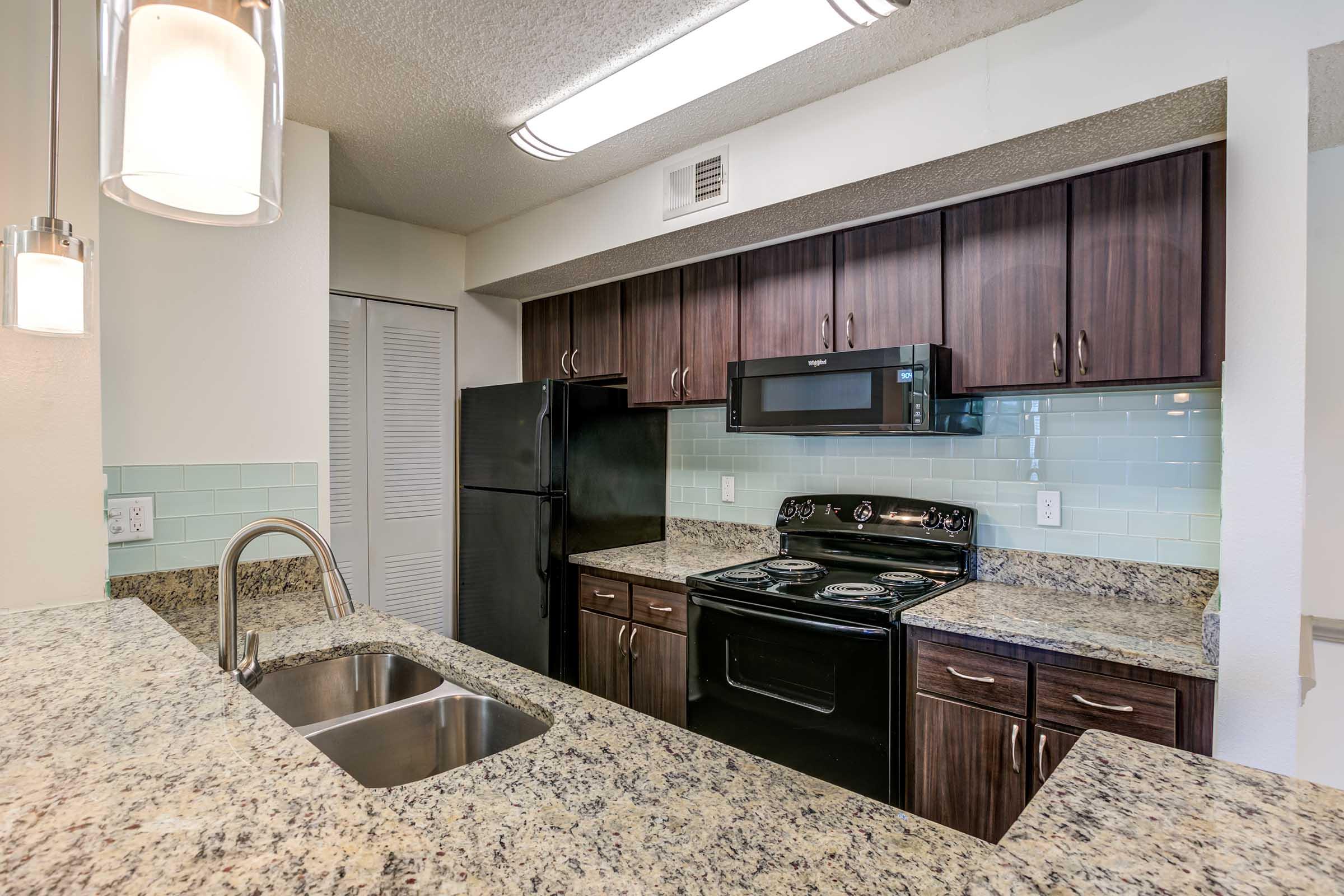 Modern kitchen featuring dark wood cabinetry, granite countertops, a black refrigerator, and a black stove with a microwave above it. Light teal backsplash tiles add a pop of color. The kitchen island includes a double sink, and overhead lighting provides a bright atmosphere.