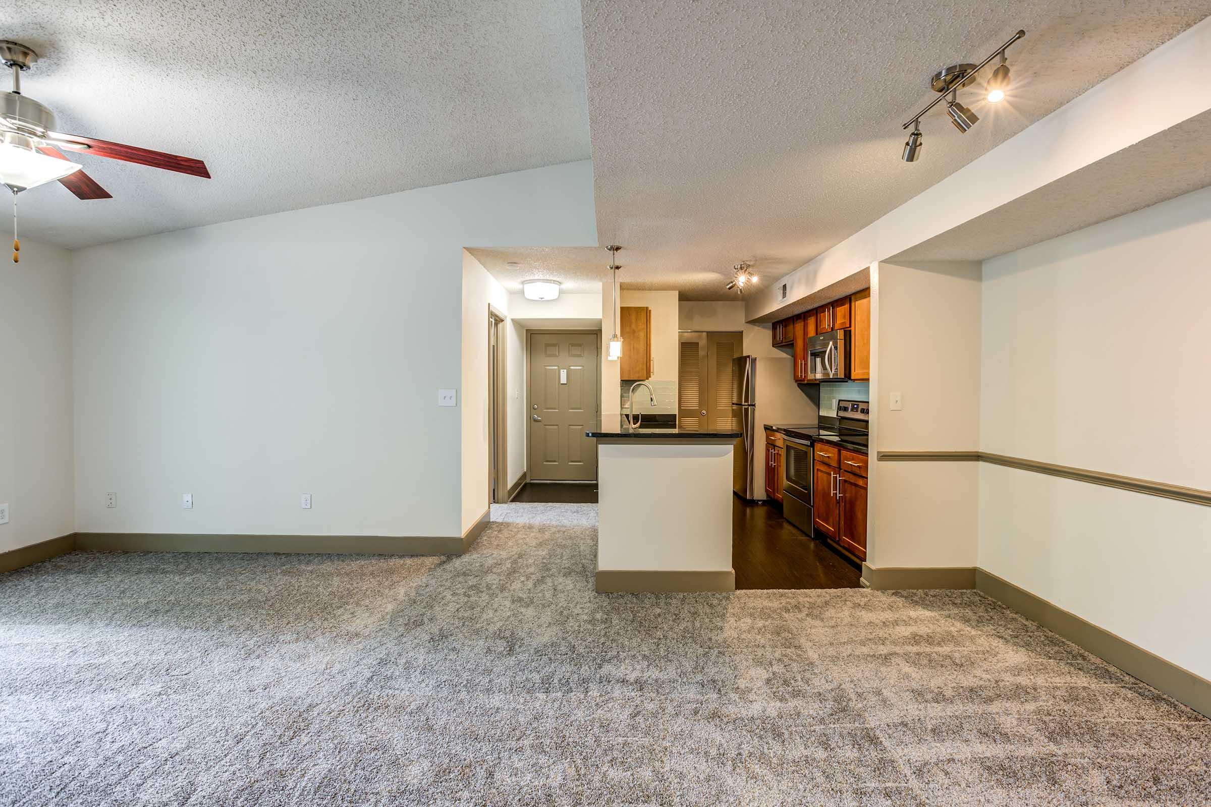 A spacious living area with a ceiling fan, carpeted flooring, and an open layout. The kitchen features stainless steel appliances and wooden cabinets, with a bar-style counter separating it from the living space. Natural light comes in from a nearby window, enhancing the bright atmosphere.