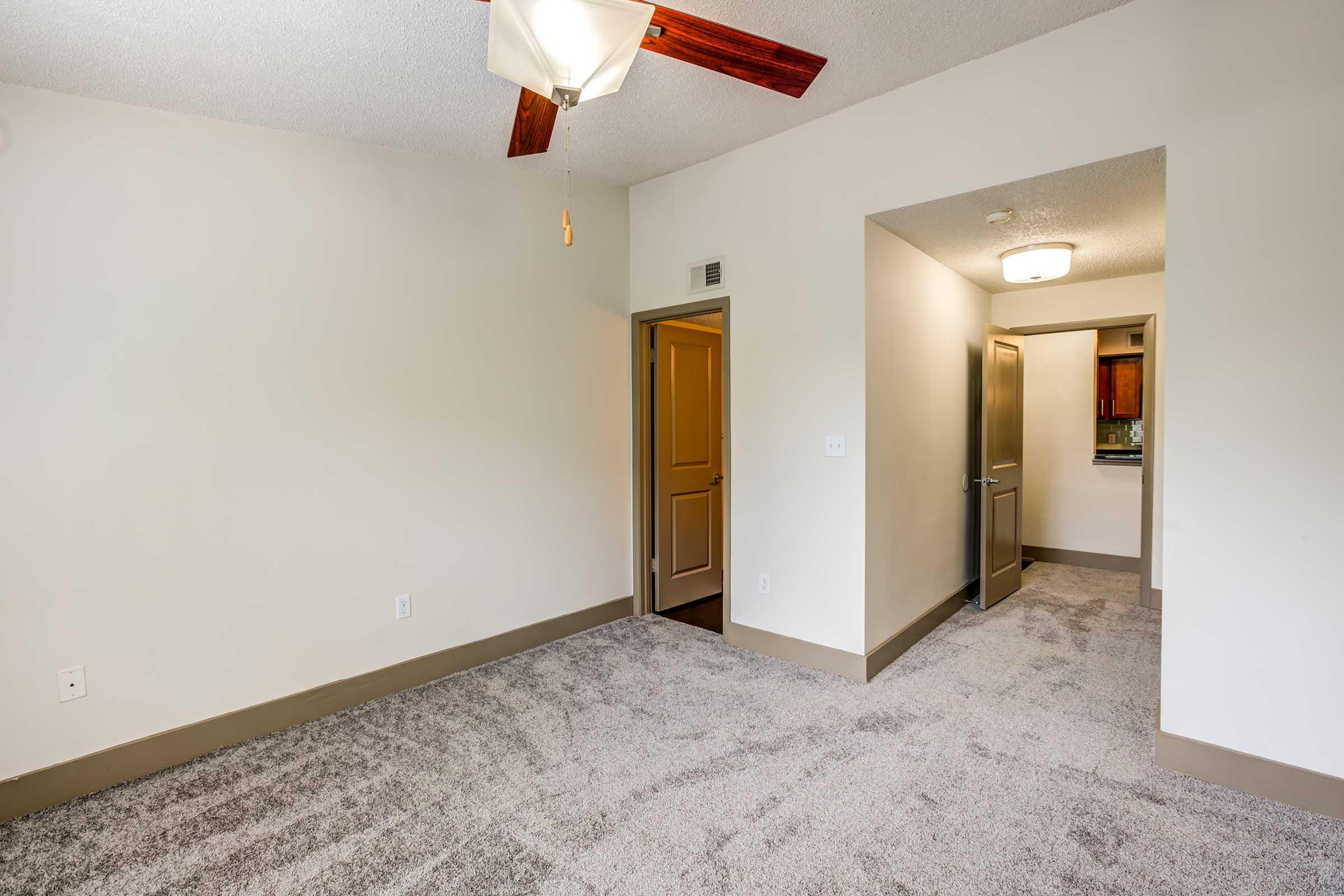 A spacious room with light-colored, freshly painted walls and plush carpet. A ceiling fan hangs from the ceiling, and there is an open doorway leading to a kitchen area in the background. The room features a warm, inviting ambiance and has a neutral color palette.