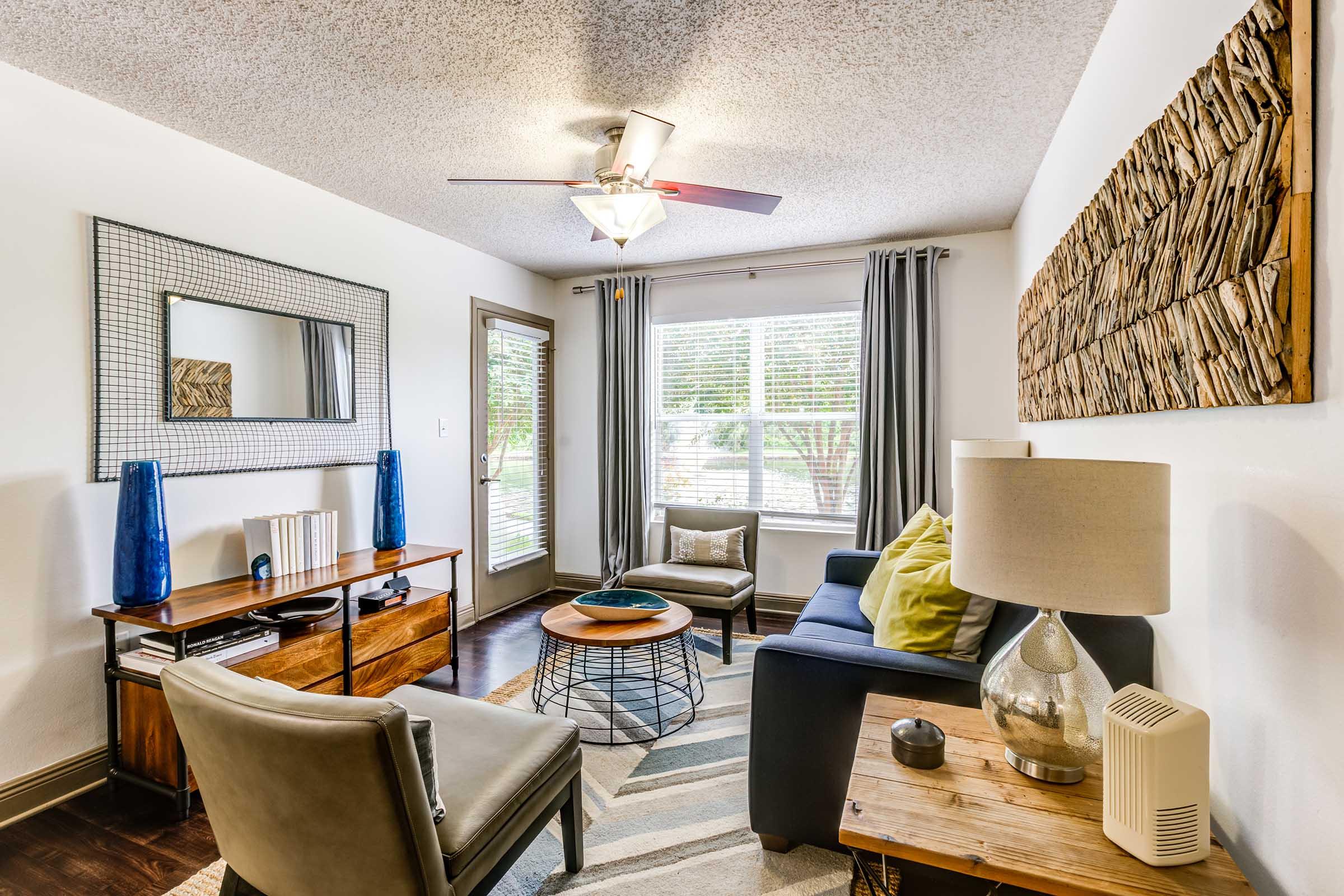 A cozy living room featuring a dark blue sofa adorned with a bright green pillow, a wooden coffee table, and an accent chair. A textured wall art piece hangs above a wooden console table with decorative items. Natural light streams in through large windows, complemented by gray curtains. A ceiling fan adds a modern touch.