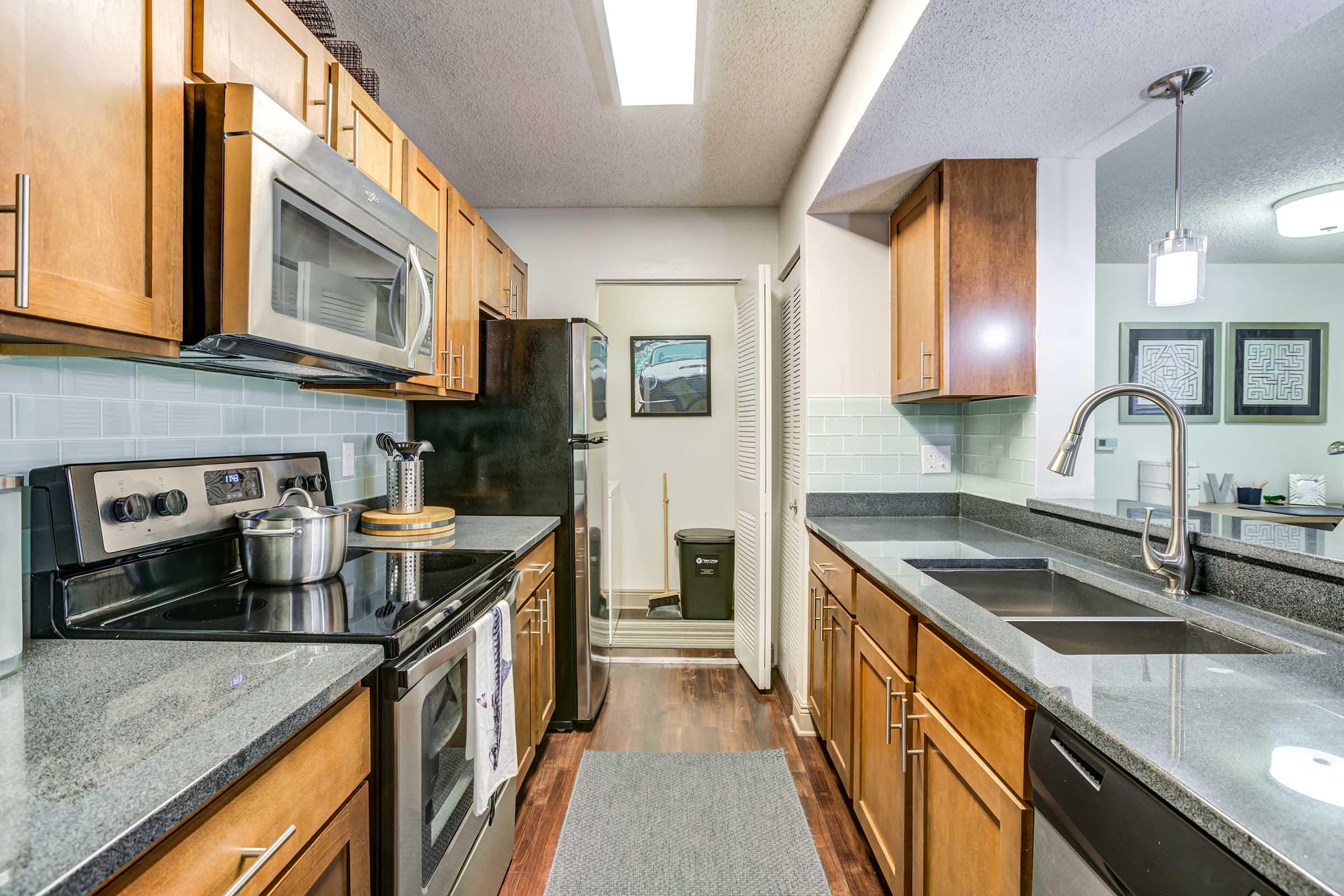 Modern kitchen featuring wooden cabinets, stainless steel appliances, and granite countertops. Includes a stove, microwave, and double sink. The layout is functional with a door leading to a storage area at the back. There is a rug on the floor and light fixtures above, creating a warm ambiance.